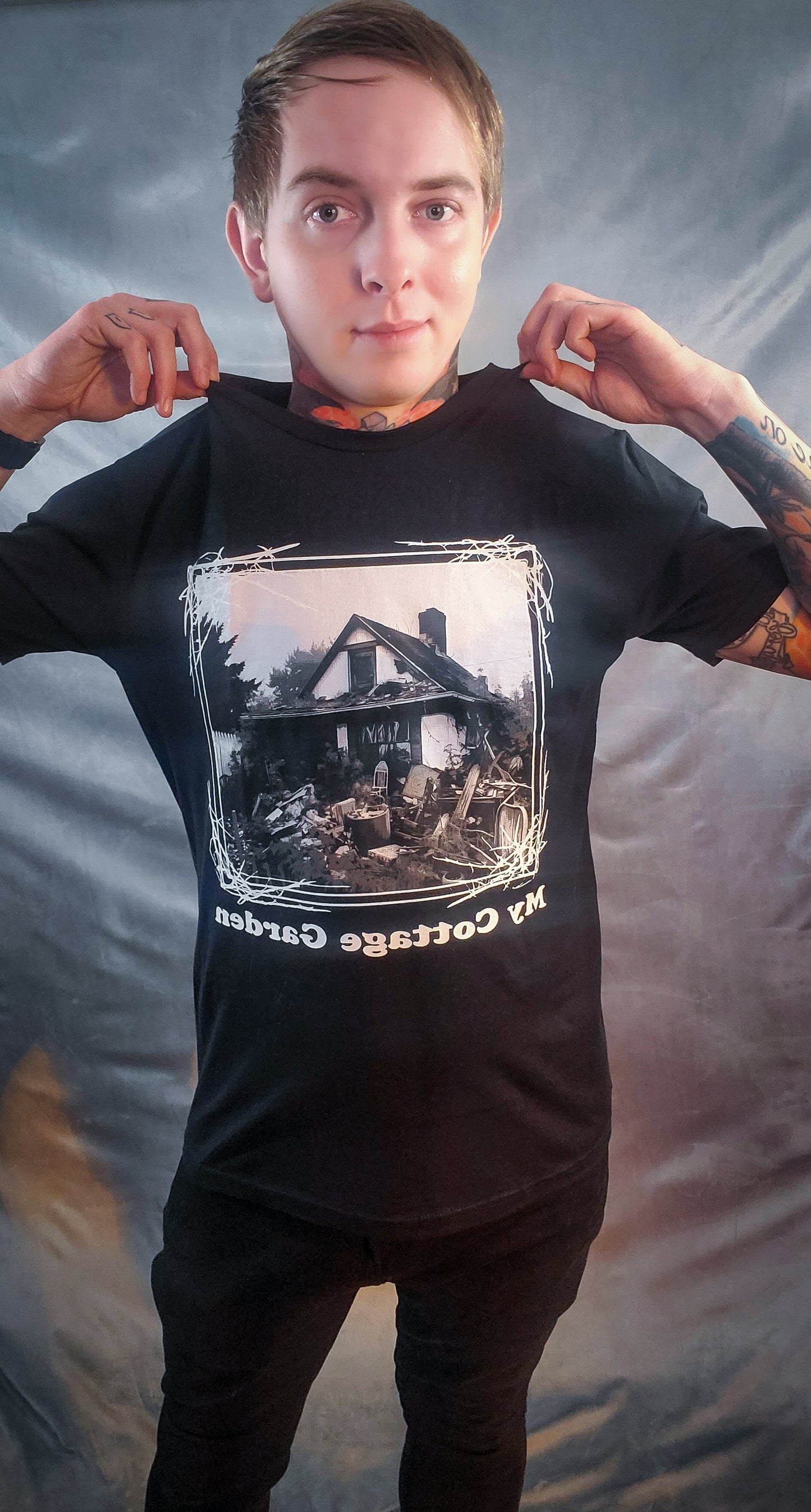 Model holding up a black tee showing a cluttered yard and house ruin print with 'My cottage Garden' script, depicting a twist on homestead life.