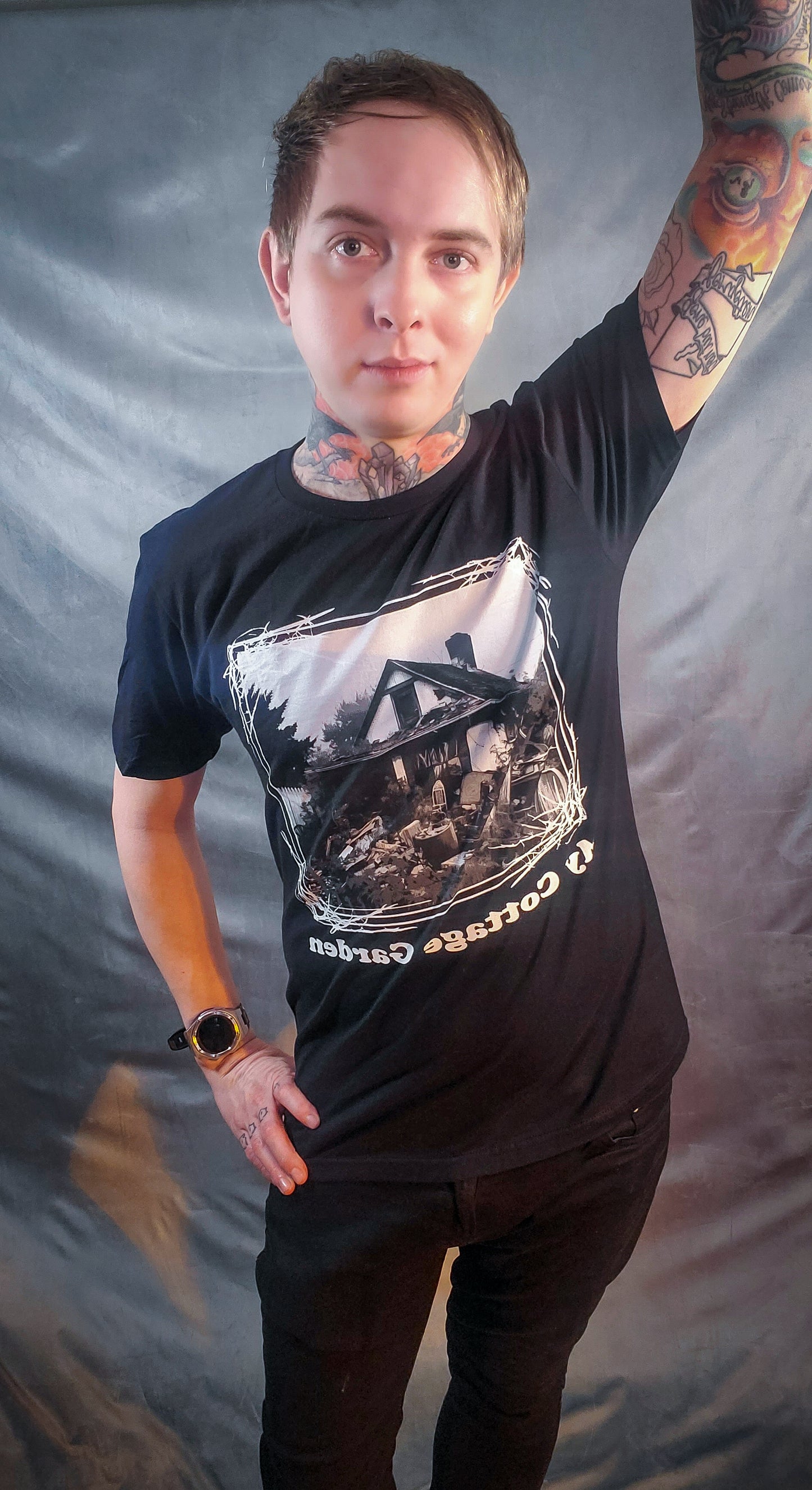 Individual posing with raised arm in a black shirt with a 'My cottage Garden' motif, showcasing a chaotic junkyard home scene.