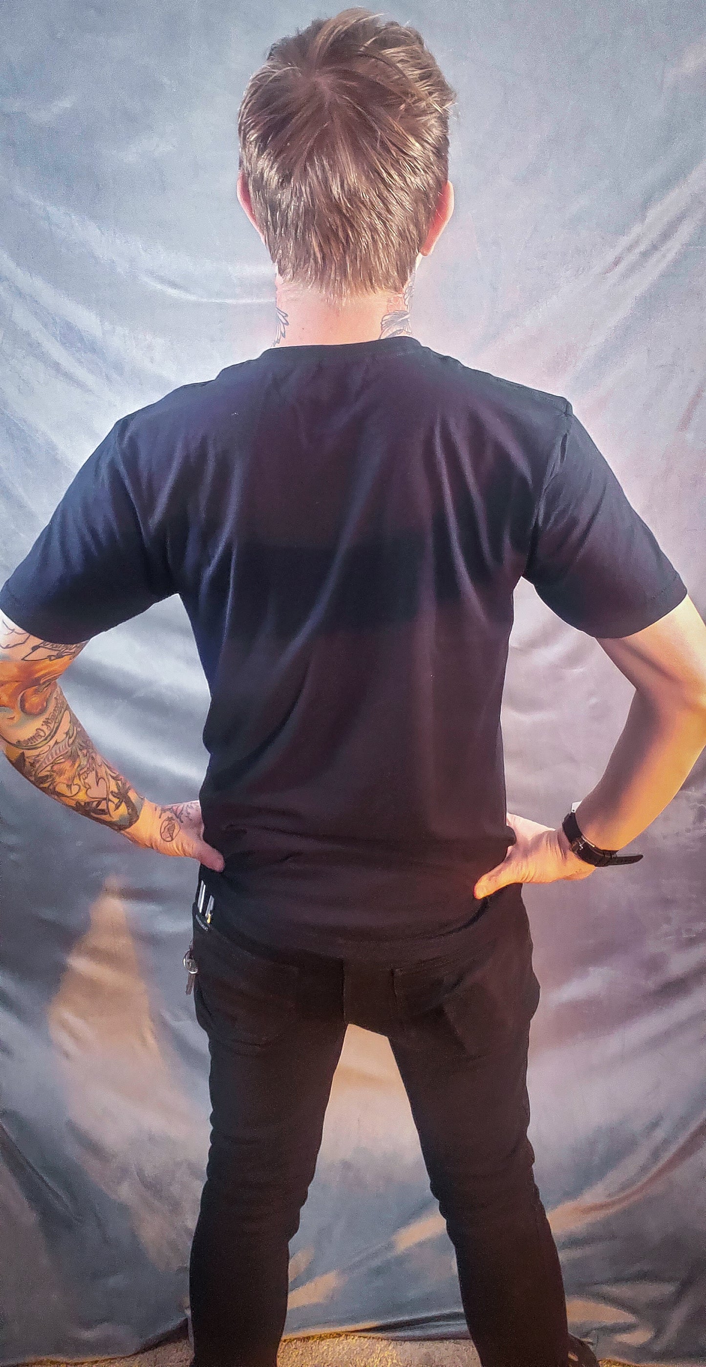Rear view of a person wearing a plain black t-shirt and black jeans, hands on hips, against a grey backdrop