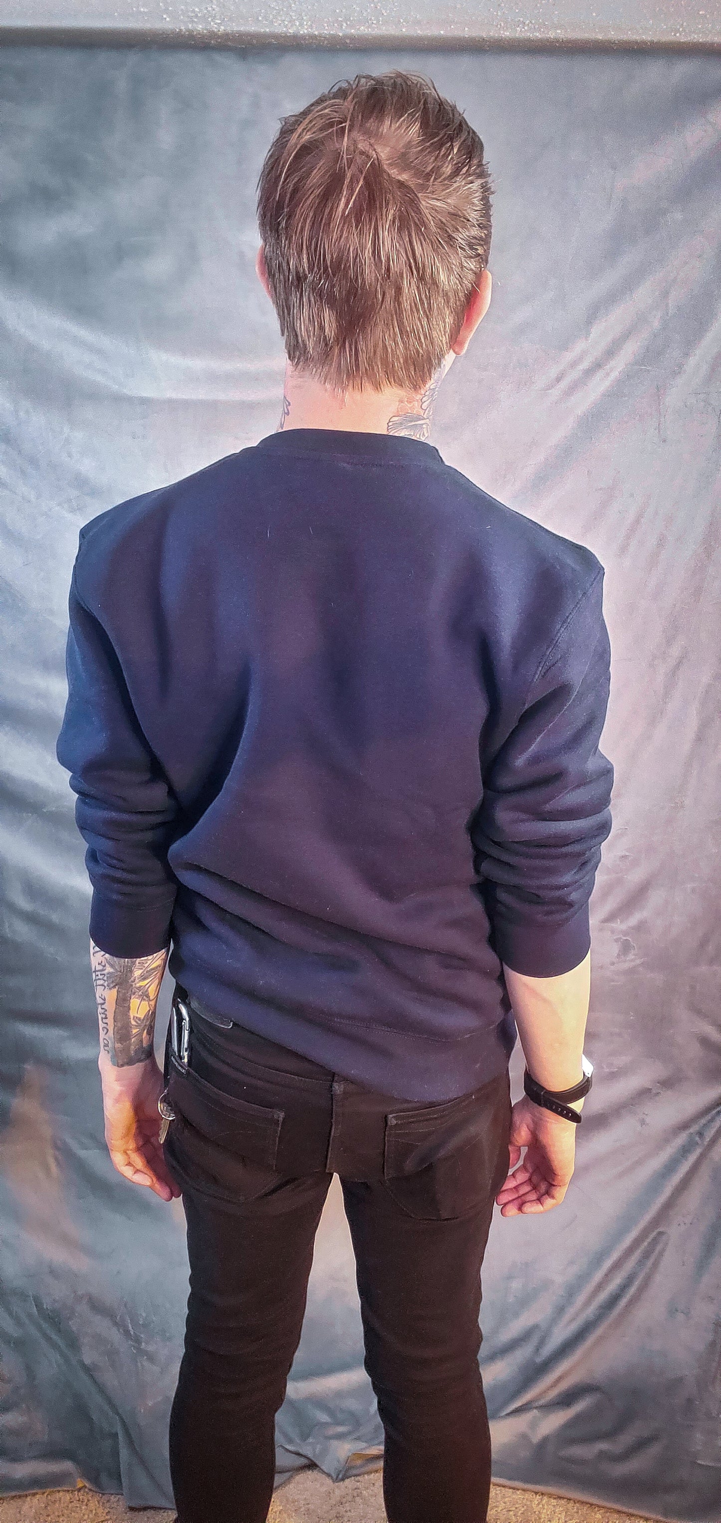 Rear view of an individual in a navy sweatshirt, hands on hips, against a textured grey background.