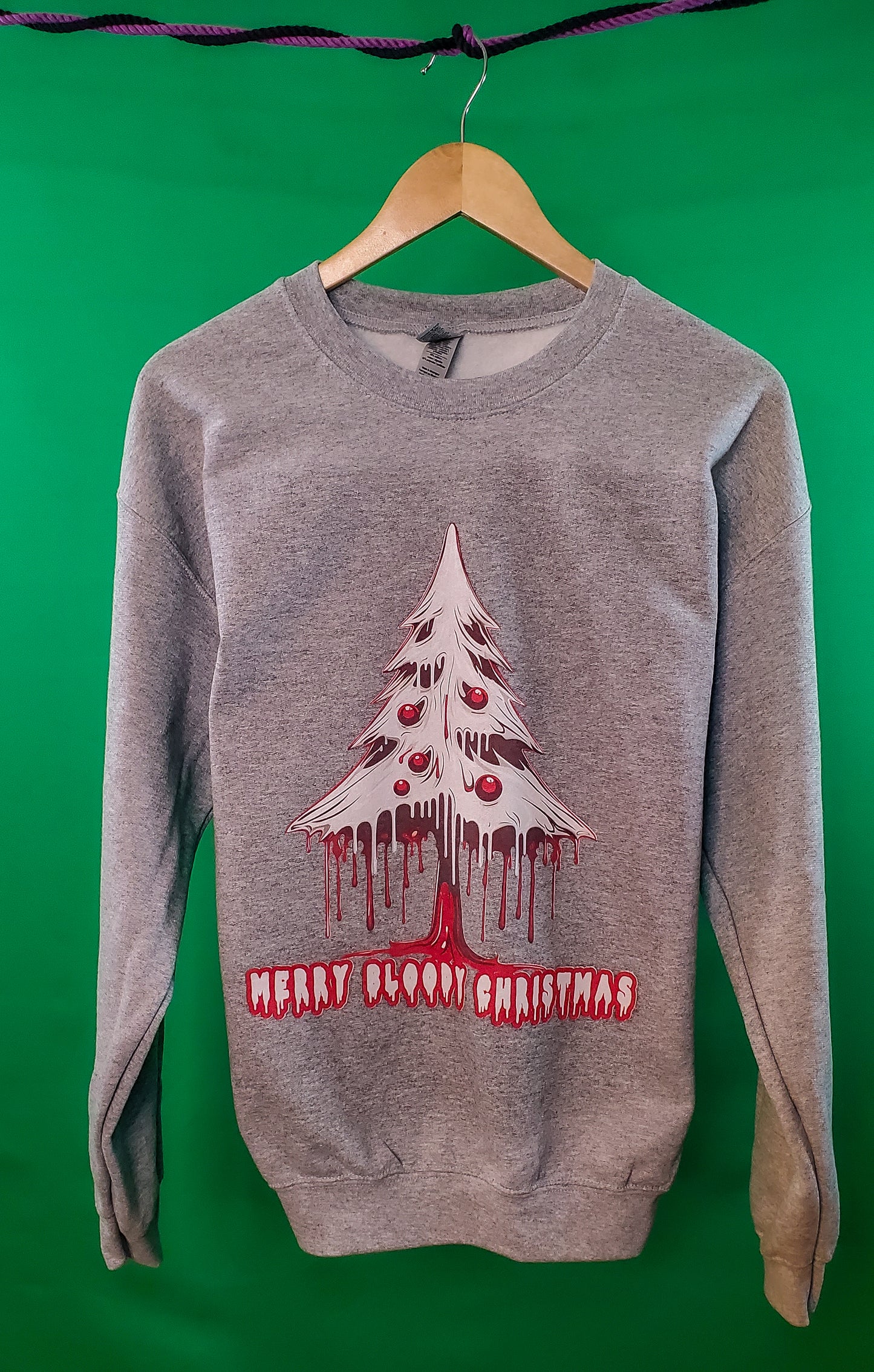 Gray crewneck sweater with a dripping pine tree design and 'Merry Bloody Christmas' in horror-themed red font, on a green background