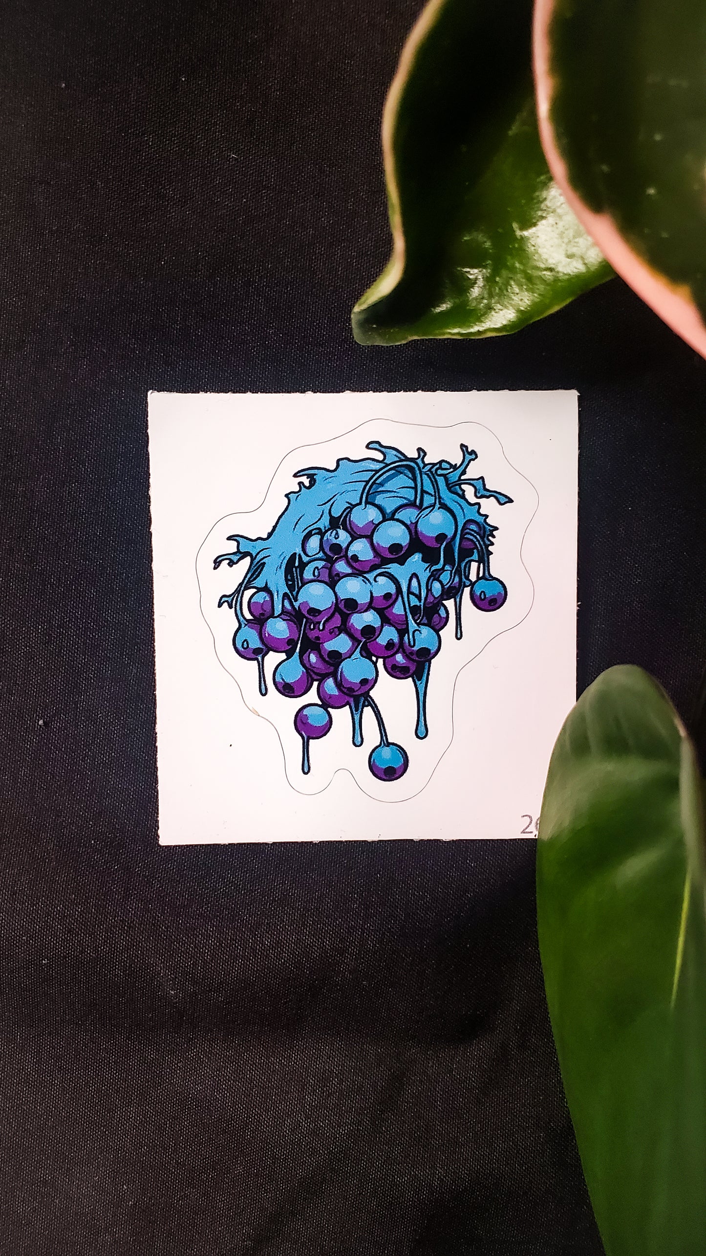 Image of a vibrant sticker featuring a surreal illustration of a dripping grape cluster in shades of blue and purple, placed against a black fabric background and surrounded by lush green leaves.