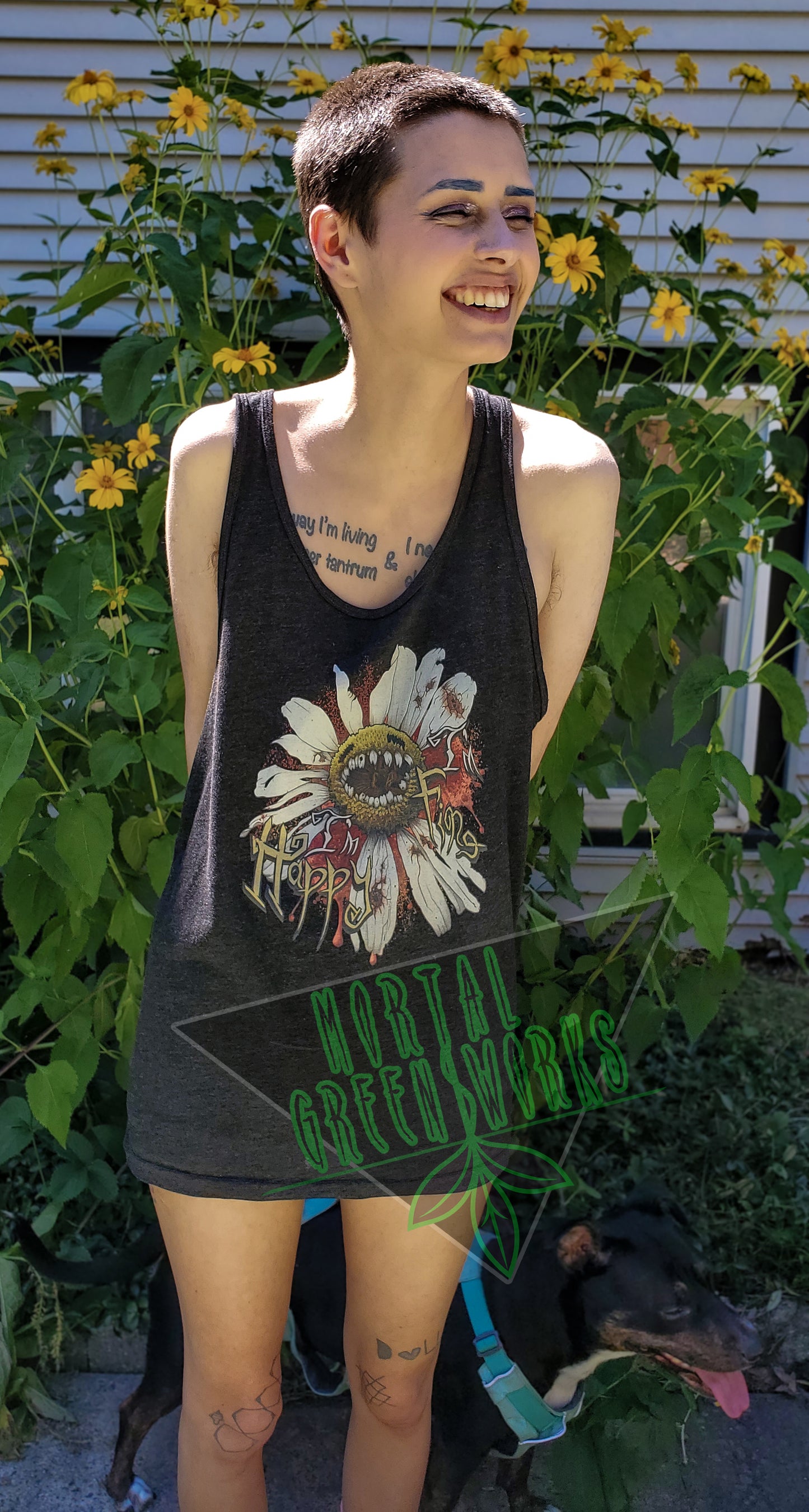 Person with short hair smiles while wearing a black tank top featuring a "Diseased Daisy" graphic with the text "I'm Happy I'm Fine." They stand in front of yellow flowers and green foliage.
