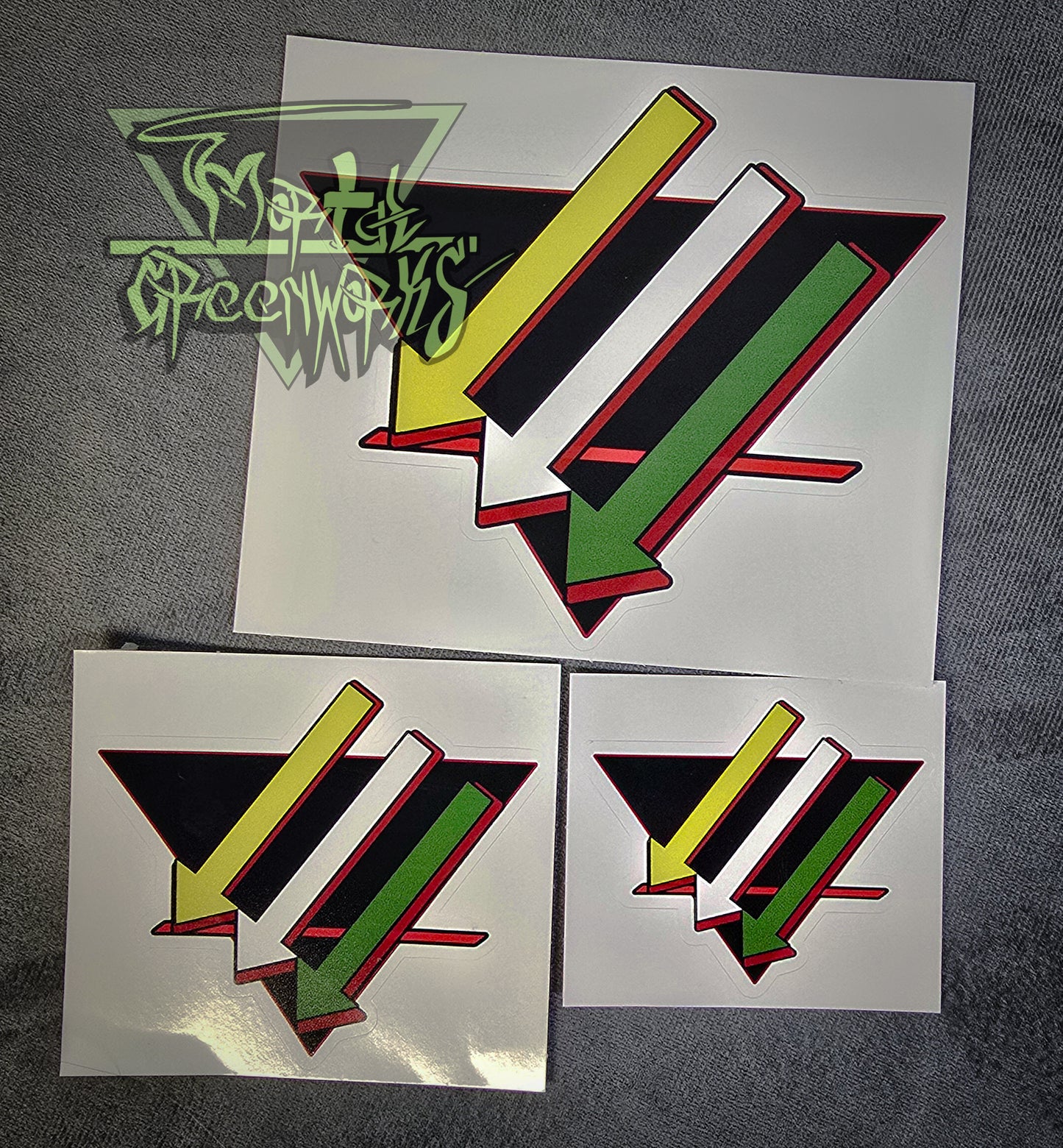 A photograph of three stickers, from large to small, on a grey tablecloth. Three arrows point down and to the left, parallel to each other, the first one gold, the second silver, and the third green. They lay over a black, upside-down triangle with a bold red line in the bottom third. The sticker has a red outline. The Mortal Greenworks watermark is in the top-left corner of the image.