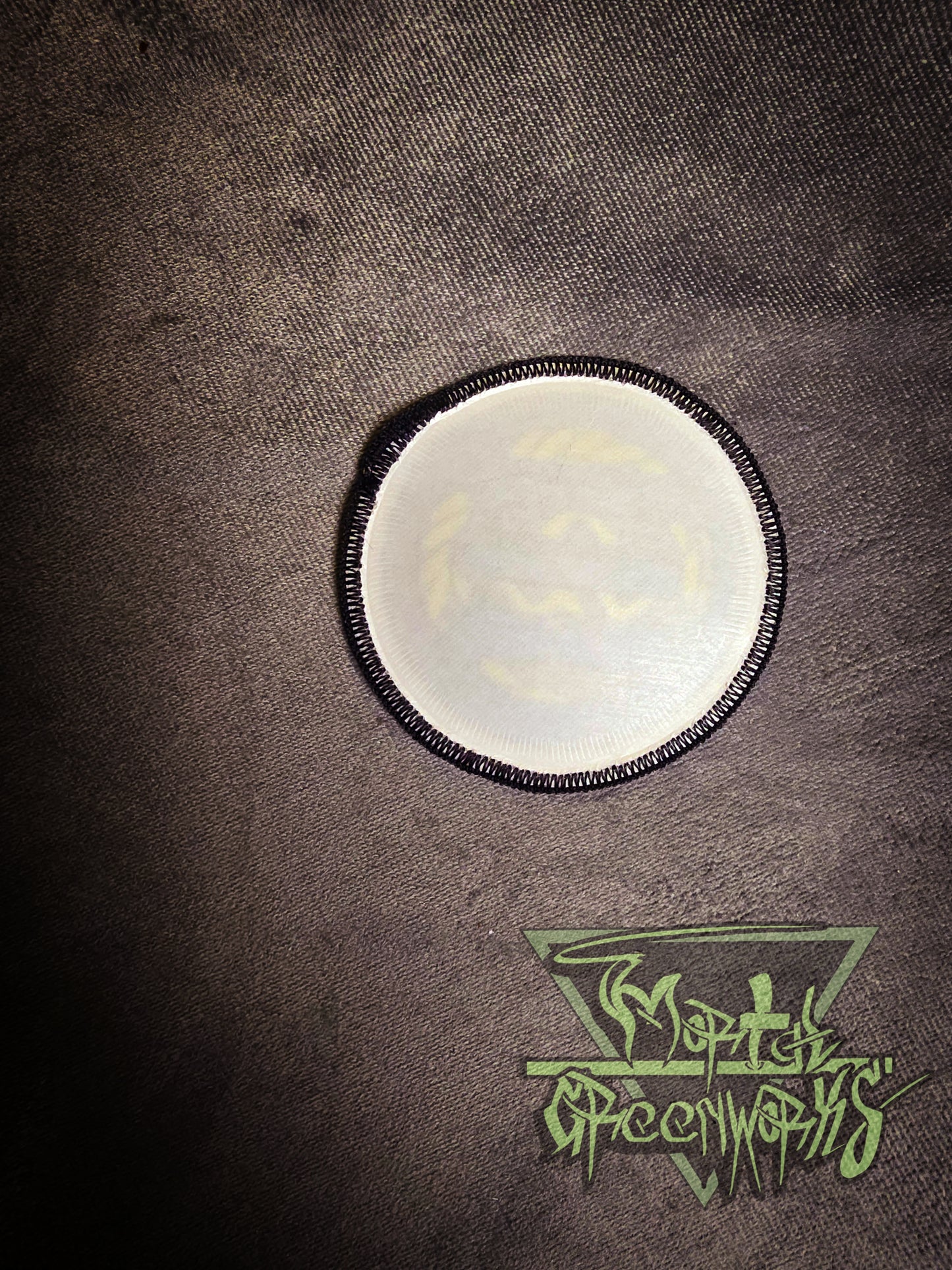 The backside of a round white iron-on patch with a black border on a textured brown surface, featuring a logo at the bottom.