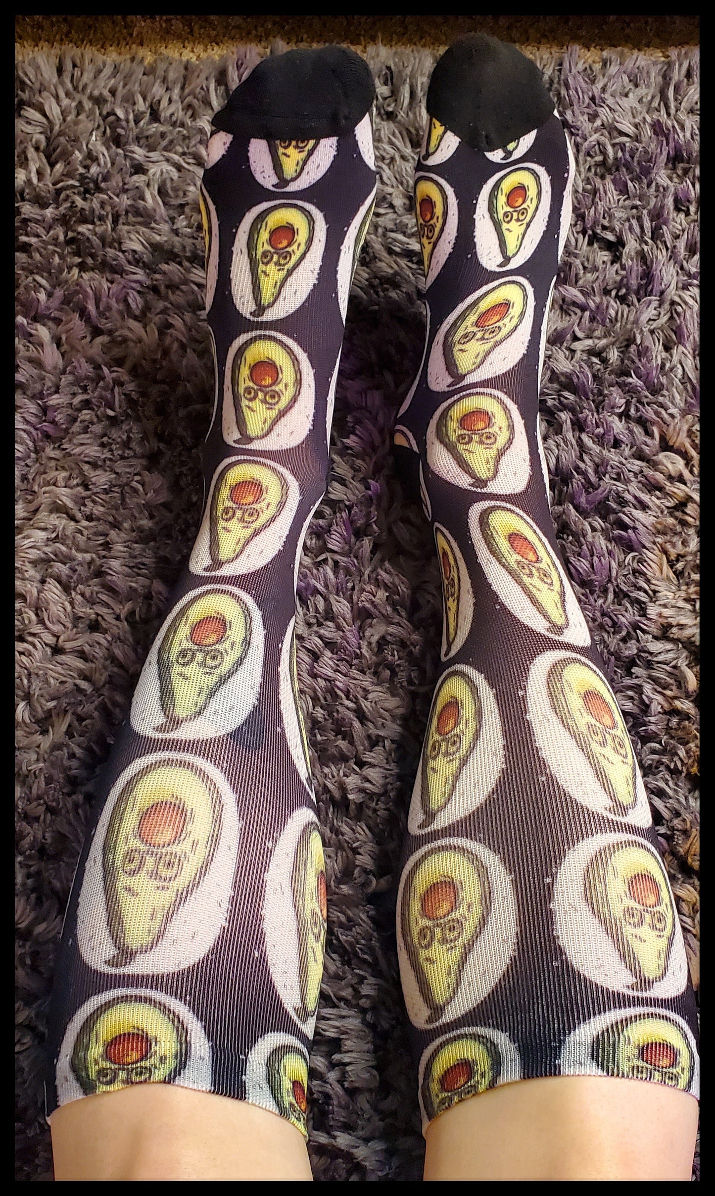 A pair of knee-high socks worn over the calves, showcasing a pattern of avocados with a surprised facial expression, where the brown pit appears to be stuffed in the mouth. The avocados have green skin and are surrounded by an off-white, painted circle, giving a halo effect on the black fabric of the socks.