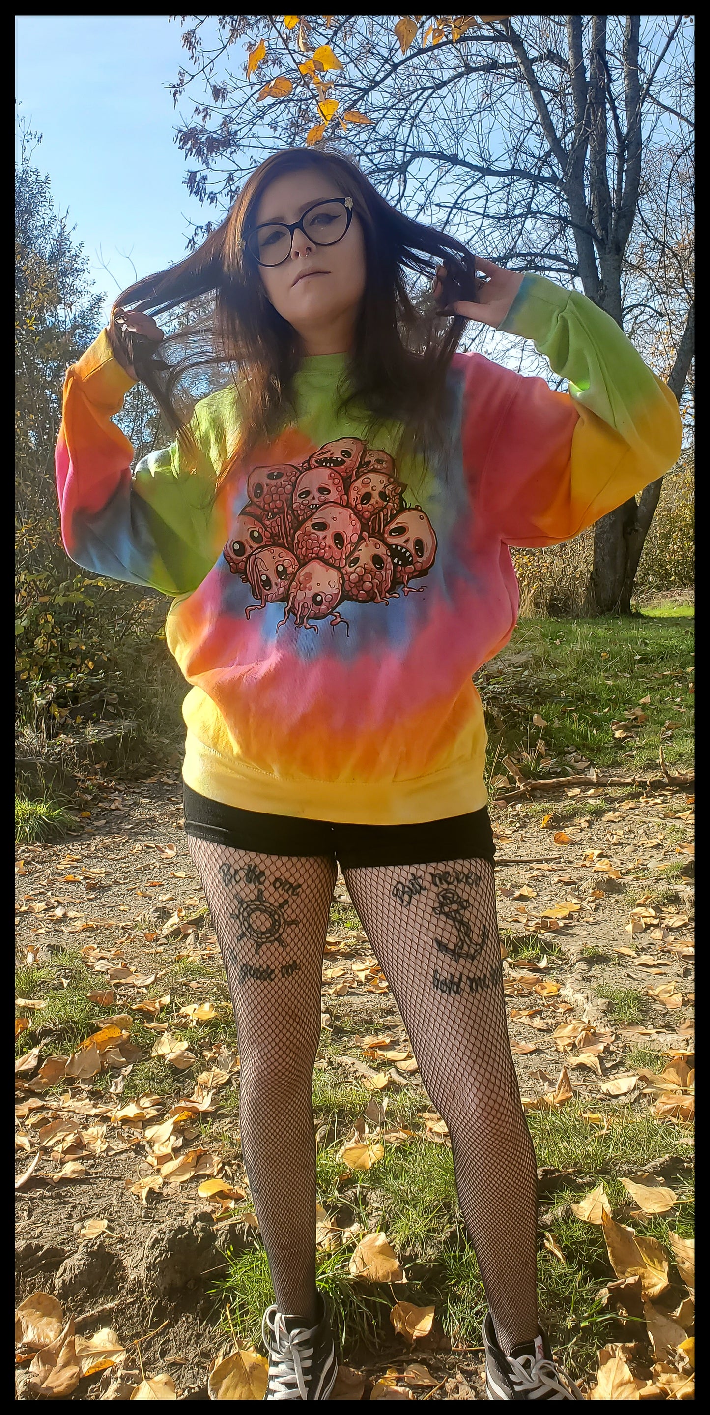 The person poses in a tie-dye sweatshirt with a unique design blending gothic and natural elements, showcasing a cluster of raspberry skulls that merge botanical and macabre aesthetics, set against the backdrop of crisp autumn leaves.