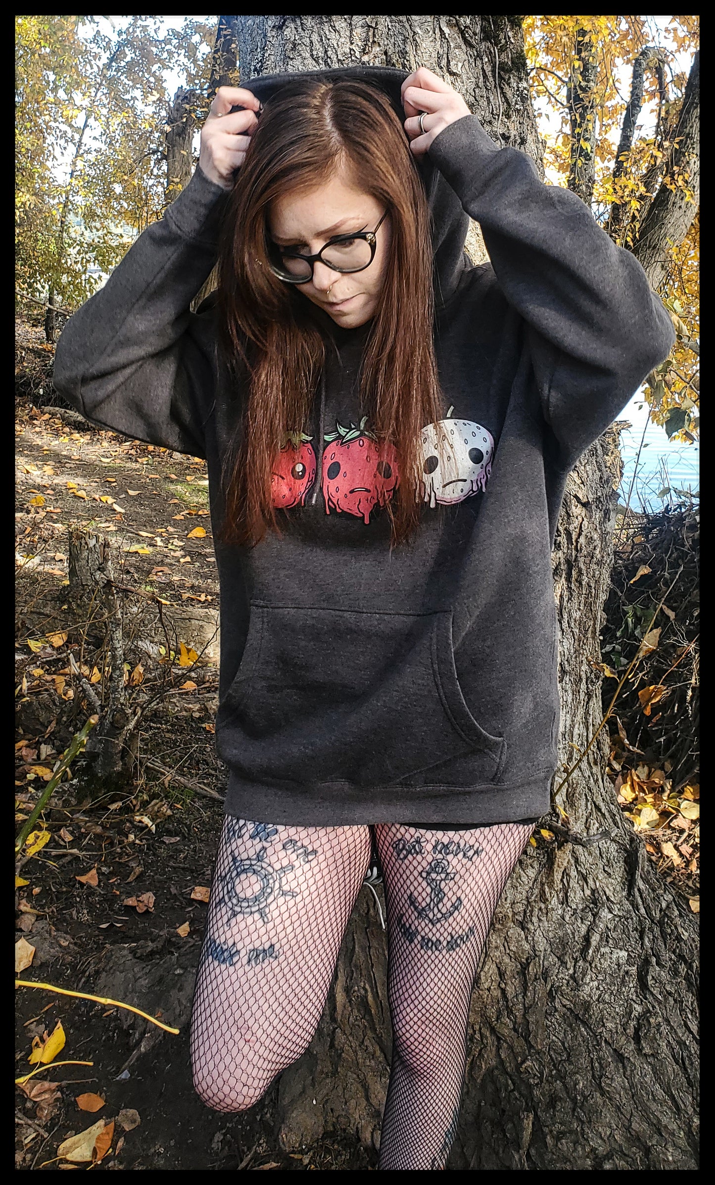 a person with glasses, seated by a tree. They're wearing a black hoodie featuring fruit designs with a horror twist, paired with fishnet tights and patterned tattoos visible on the legs. The setting includes fallen autumn leaves and a background view of water through trees, suggesting a peaceful outdoor scene.