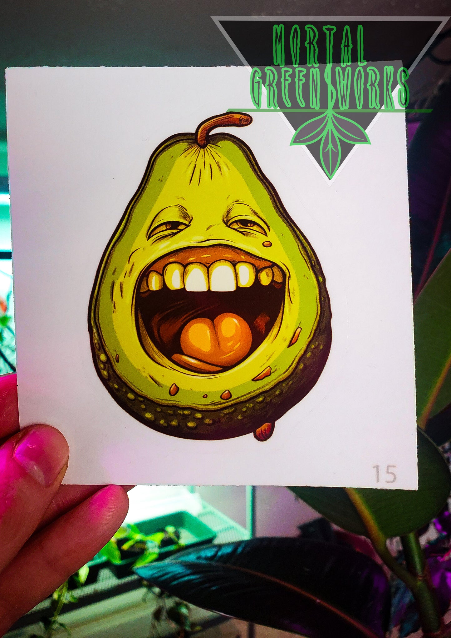 A playful sticker of an avocado with a wide, toothy smile held up against an indoor plant background, featuring 'Mortal Greenworks' logo