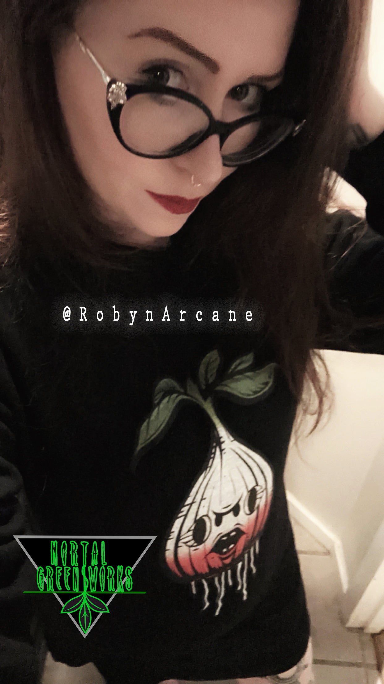 Person in a black sweatshirt featuring a cartoonish, sinister radish design, under the Mortal Greenworks logo, tagged with @RobynArcane.