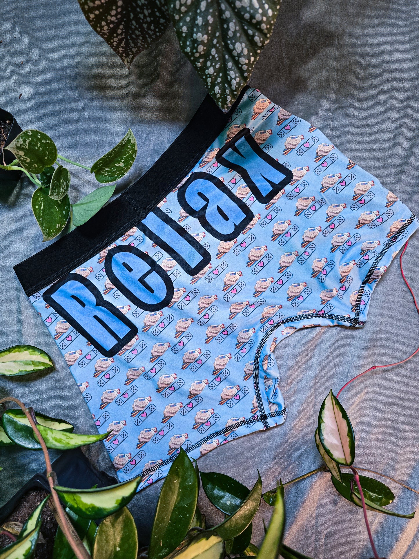 Boxer briefs displayed with the back side facing up, featuring bold blue text that reads 'Relax' over the chicken and bandaid pattern, presented against a gray backdrop with surrounding green plants.