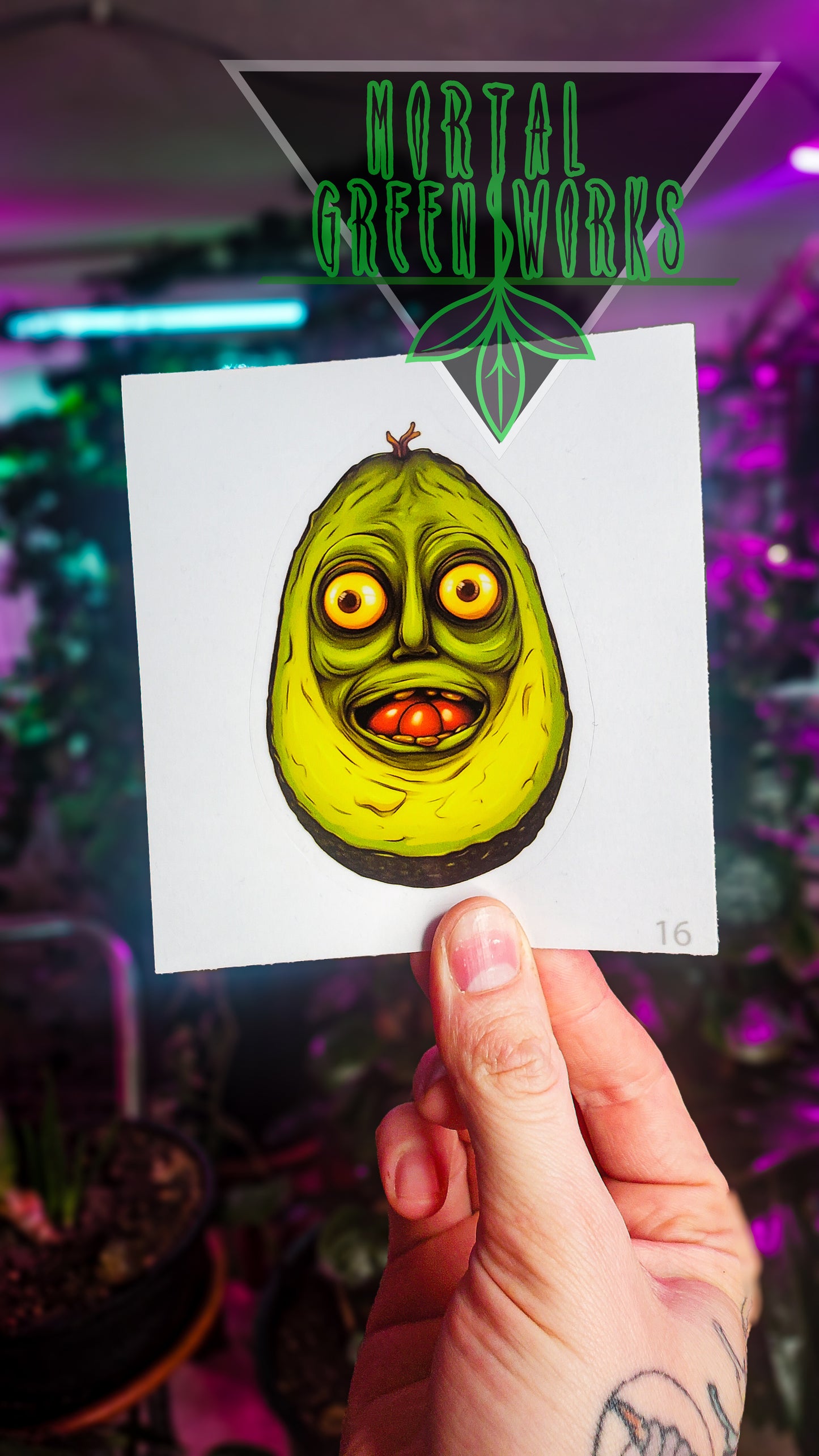 A sticker of an avocado with a comically alarmed face, held by a hand with 'Mortal Greenworks' logo above.