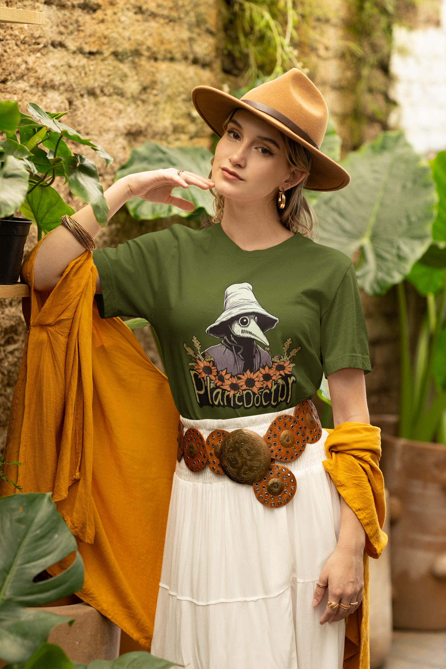 Stylish woman in a boho-chic outfit with a military green 'Plant Doctor' t-shirt posing amidst verdant foliage, exuding a blend of nature and fashion.