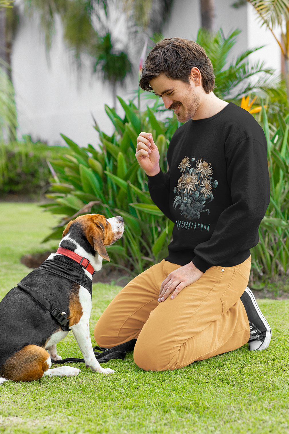 Man in a black sweatshirt with chrysanthemum design kneeling on grass beside a beagle dog.