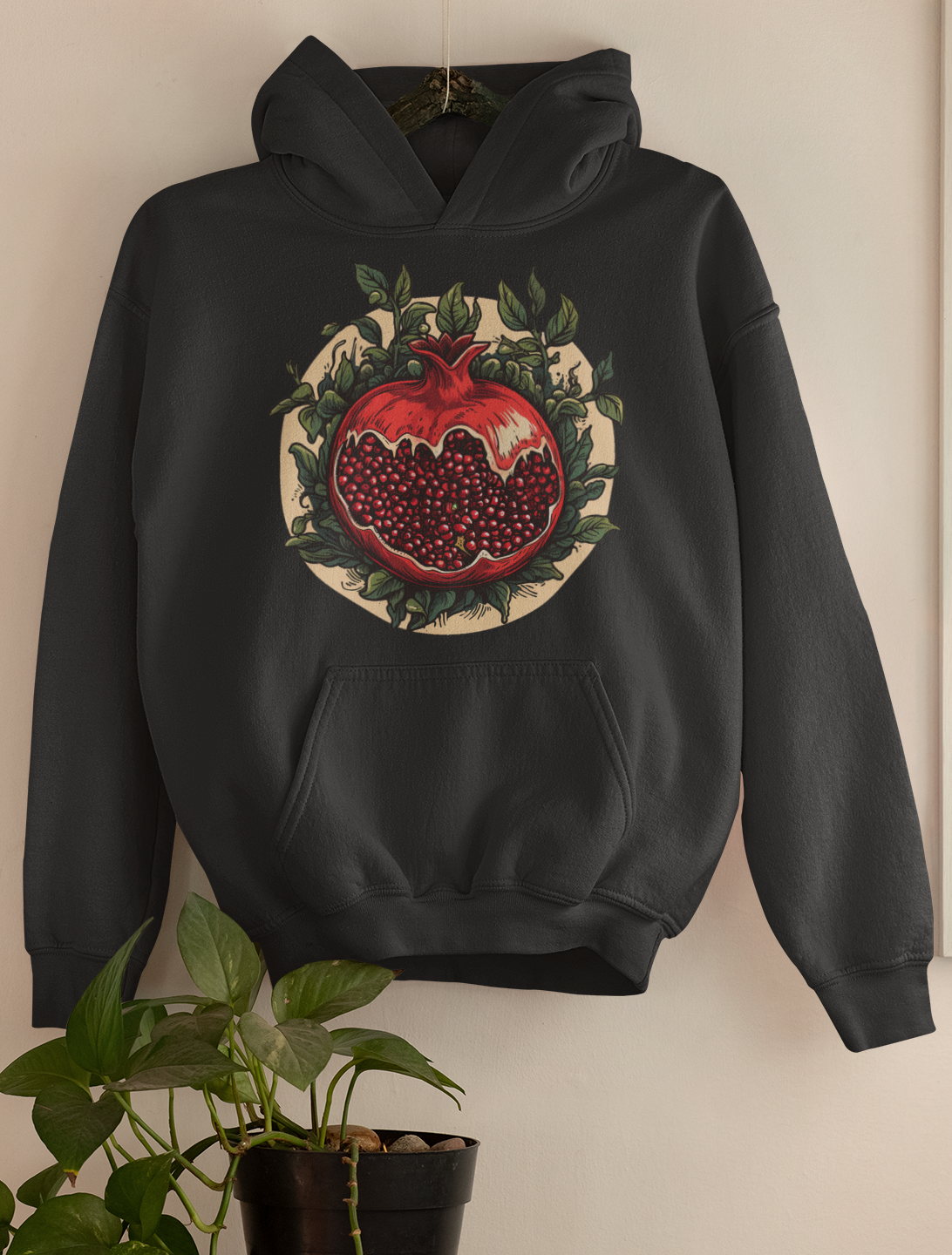 Blackhoodie featuring a detailed pomegranate illustration surrounded by foliage, hung as wall decor over a green houseplant, reflecting a darkly whimsical style.