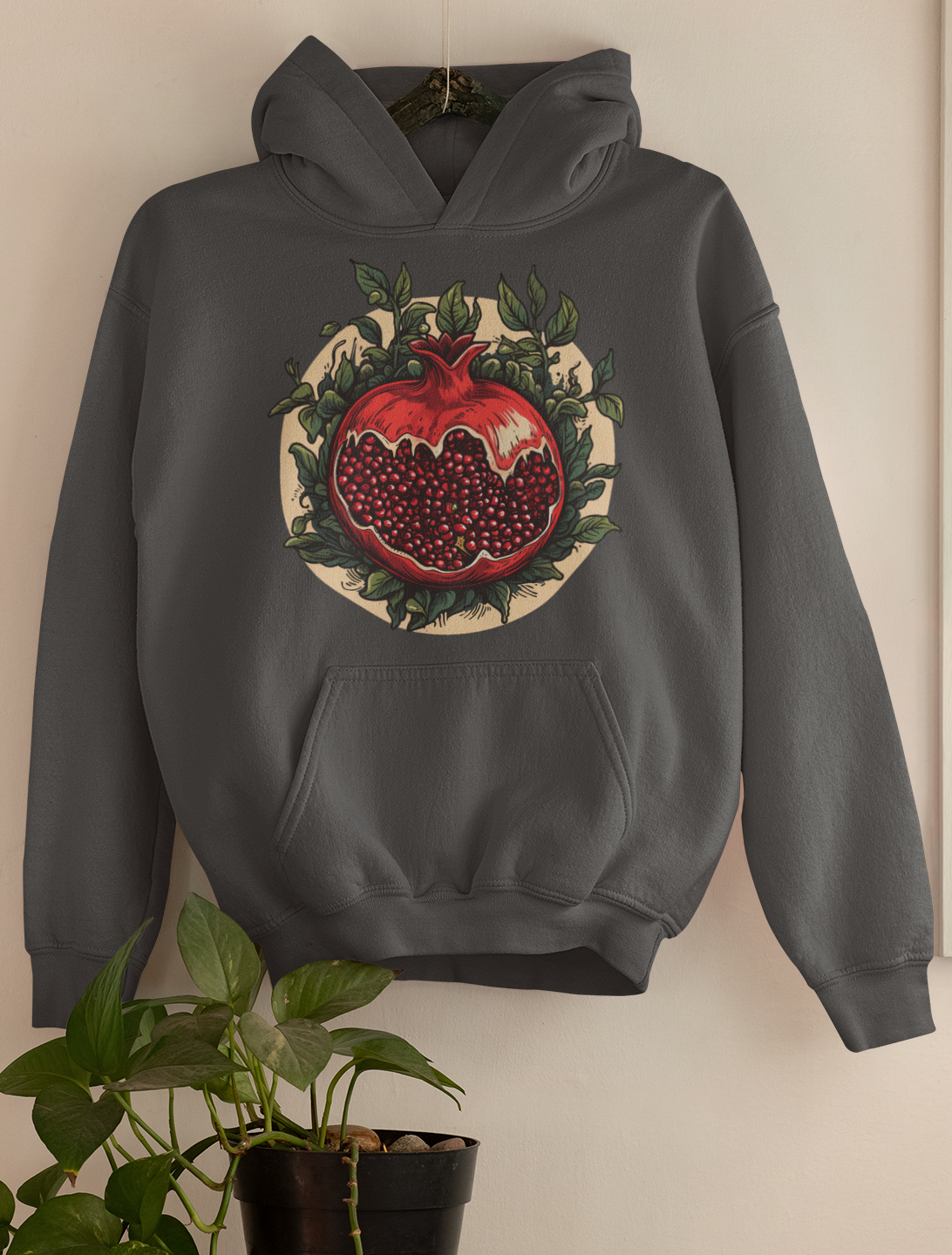 Charcoal heather hoodie featuring a detailed pomegranate illustration surrounded by foliage, hung as wall decor over a green houseplant, reflecting a darkly whimsical style.