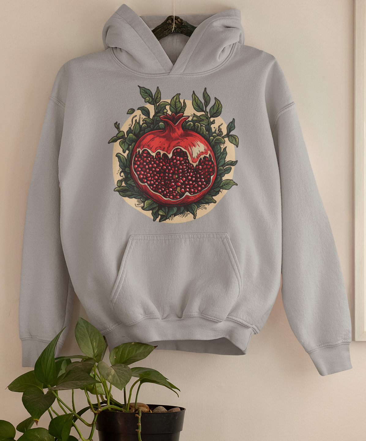 Heather grey hoodie with a vivid pomegranate design encircled by lush greenery, displayed on a wall above a potted plant, embodying a cottagecore horror aesthetic.