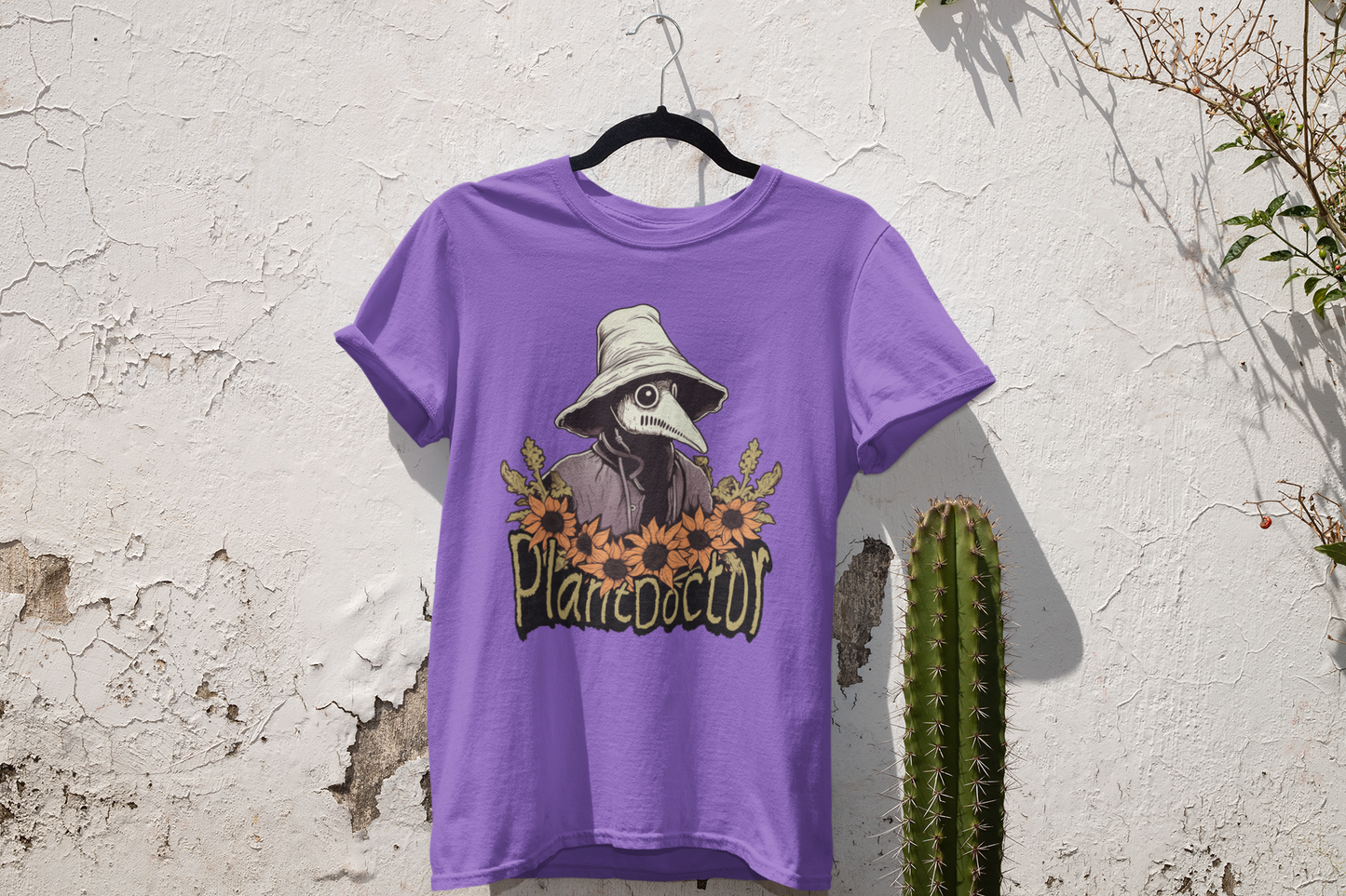 Heather team purple t-shirt with a Plague Doctor as a Plant Doctor design hanging on a white wall, complemented by natural greenery and a cactus, showcasing a rustic and earthy vibe.