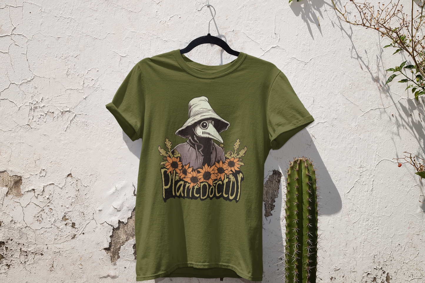 Military green t-shirt with a 'Plant Doctor' design hanging on a white wall, complemented by natural greenery and a cactus, showcasing a rustic and earthy vibe.