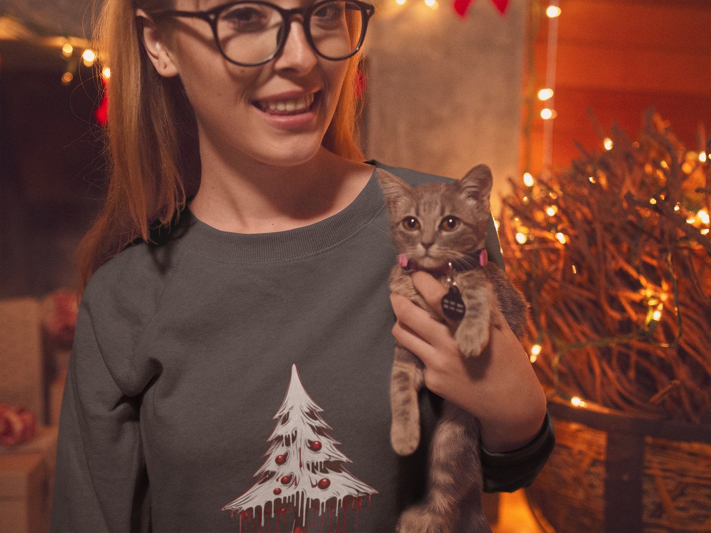 Woman in glasses holding a kitten, wearing a grey sweatshirt with a "Merry Bloody Christmas" tree design.