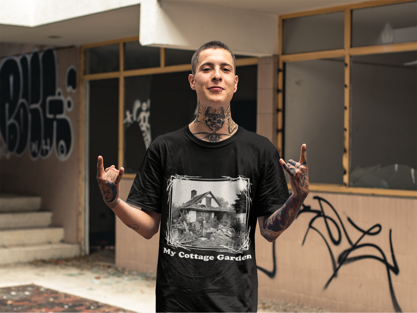 Tattooed man in a black t-shirt with 'My Cottage Garden' print stands in front of an abandoned building, representing a rebellious spirit with a darkly humorous twist.