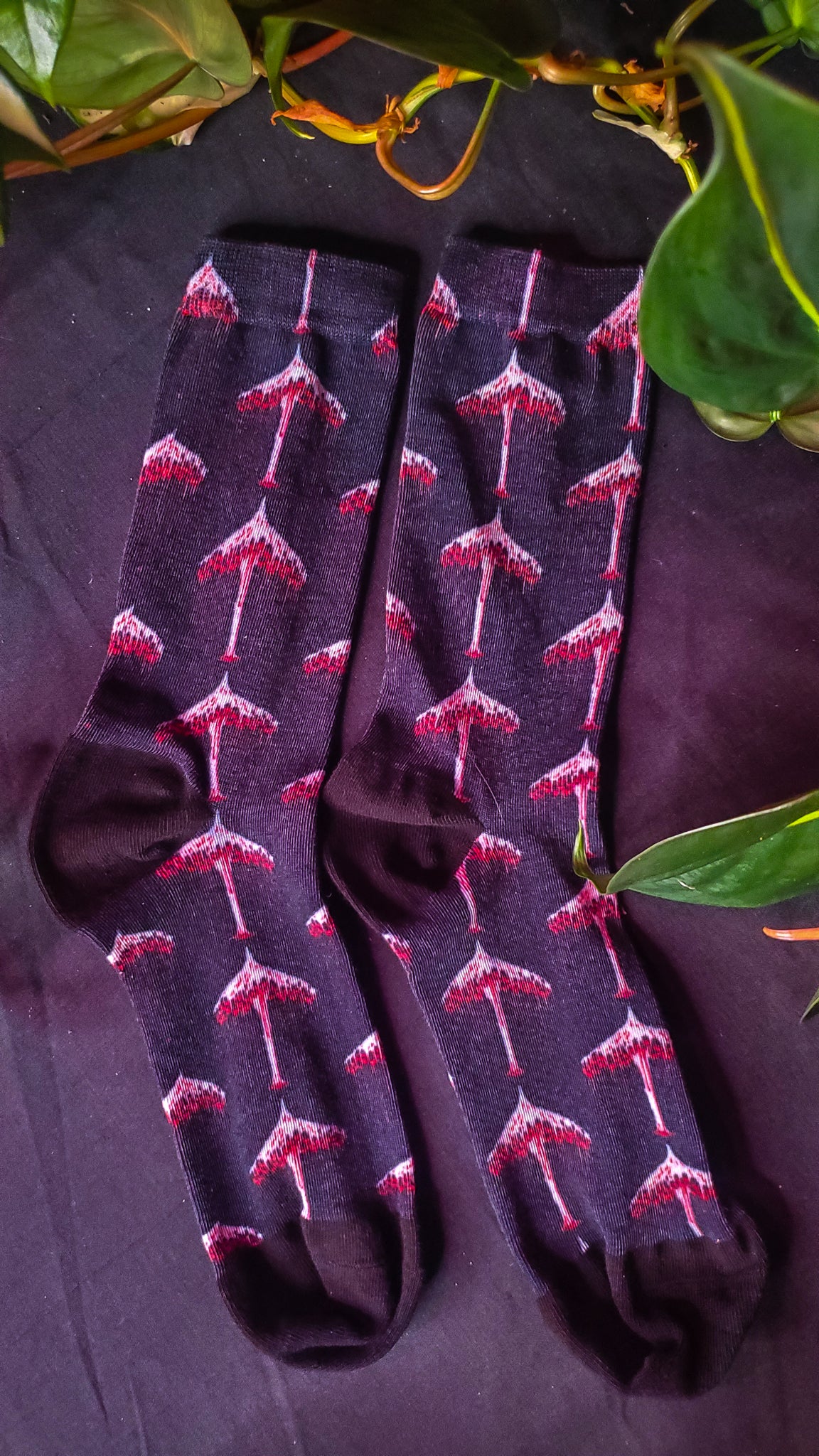 Socks with eerie bleeding mushroom patterns on a black background, nestled among green leaves.