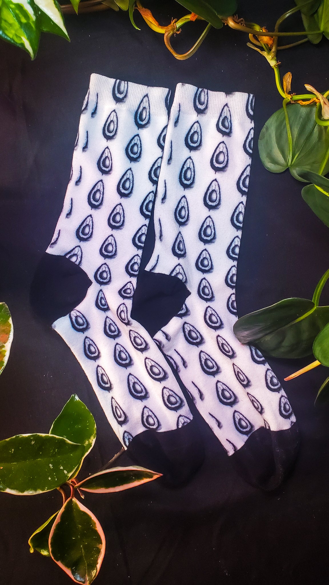 A pair of socks with an avocado pattern printed in black on a white background is displayed on a dark fabric surface. The all-seeing eye design is scattered across the socks, creating a mystical and intriguing appearance.