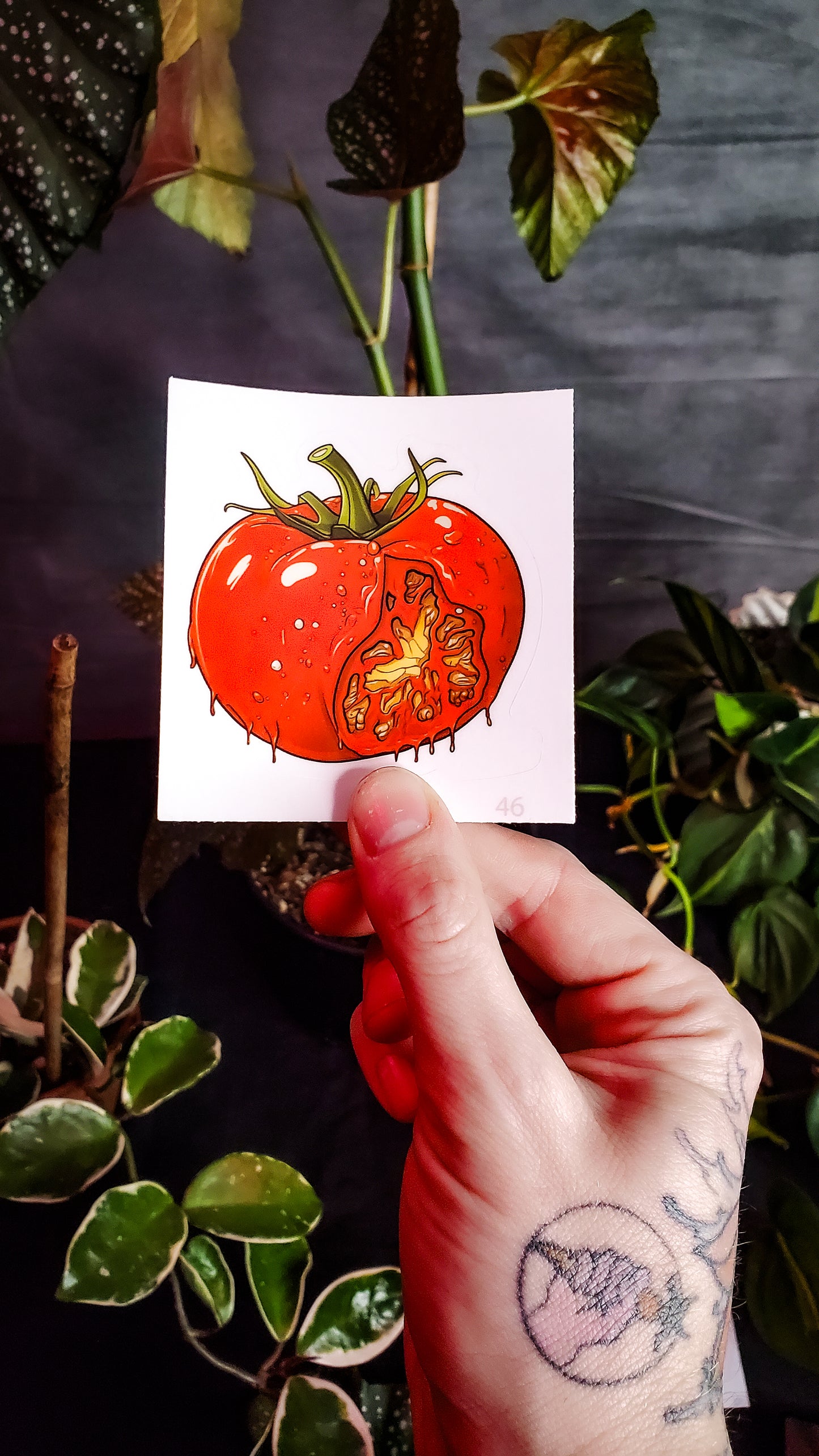 A hand holding a sticker of a ripe tomato with a scary face carved inside, amidst lush houseplants.