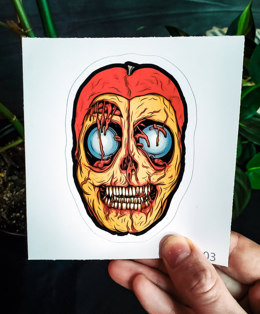 A hand holding a sticker depicting a macabre passion fruit with a skull-like interior, against greenery.