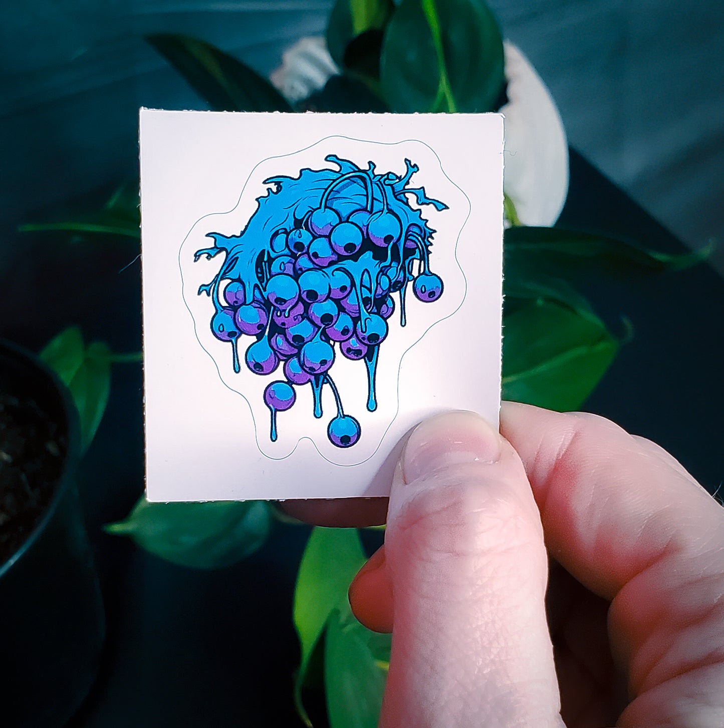 A hand holding a sticker of a dripping blueberry cluster with a blue overlay, against a backdrop of green leaves