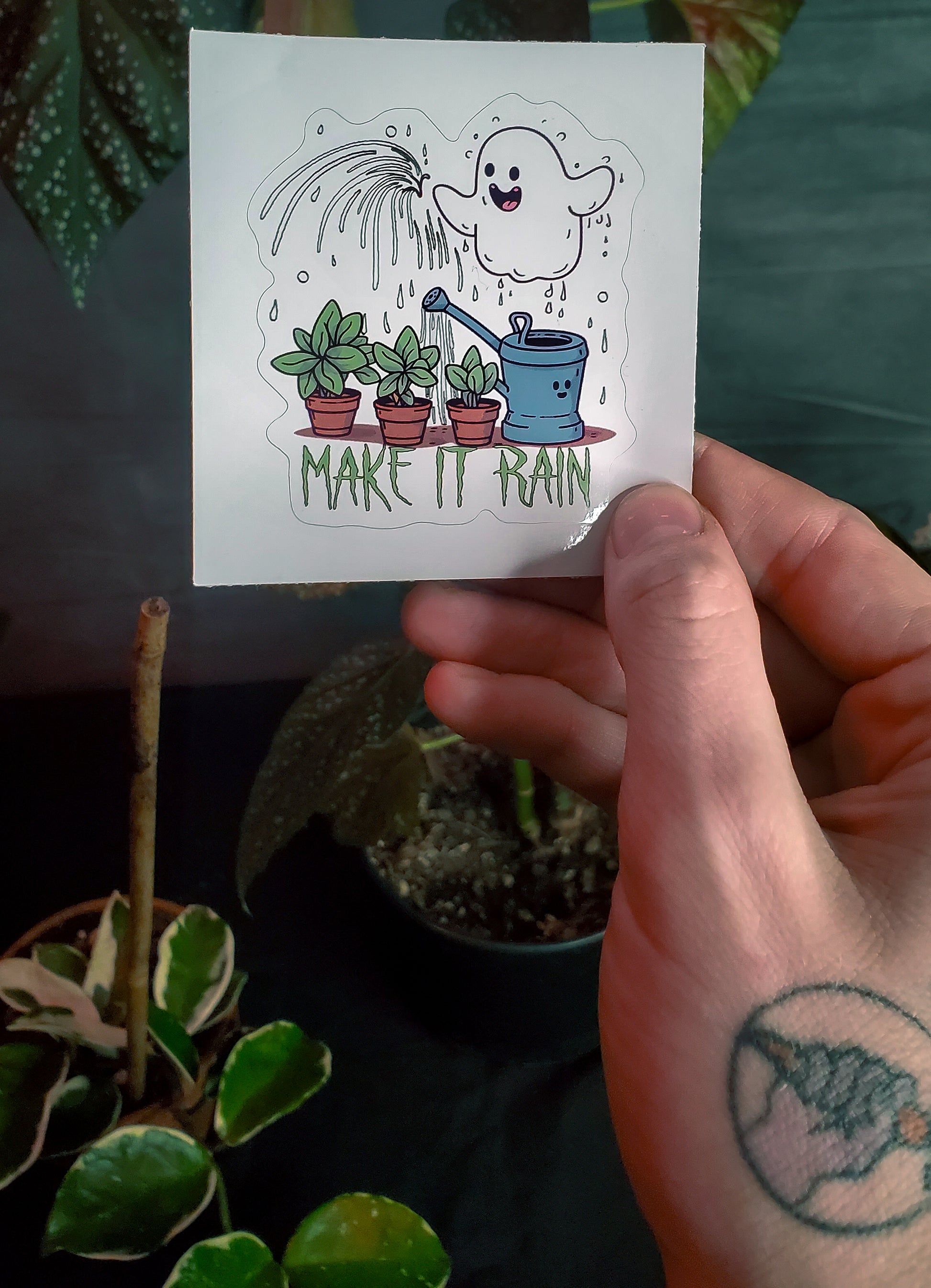 A hand holding a sticker with a cheerful ghost watering houseplants, text says 'MAKE IT RAIN'