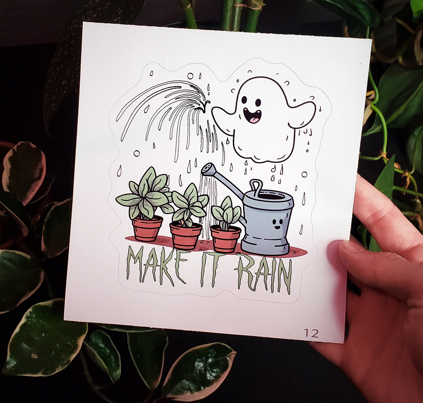 A sticker showing a happy ghost watering plants with a 'MAKE IT RAIN' slogan, held against foliage