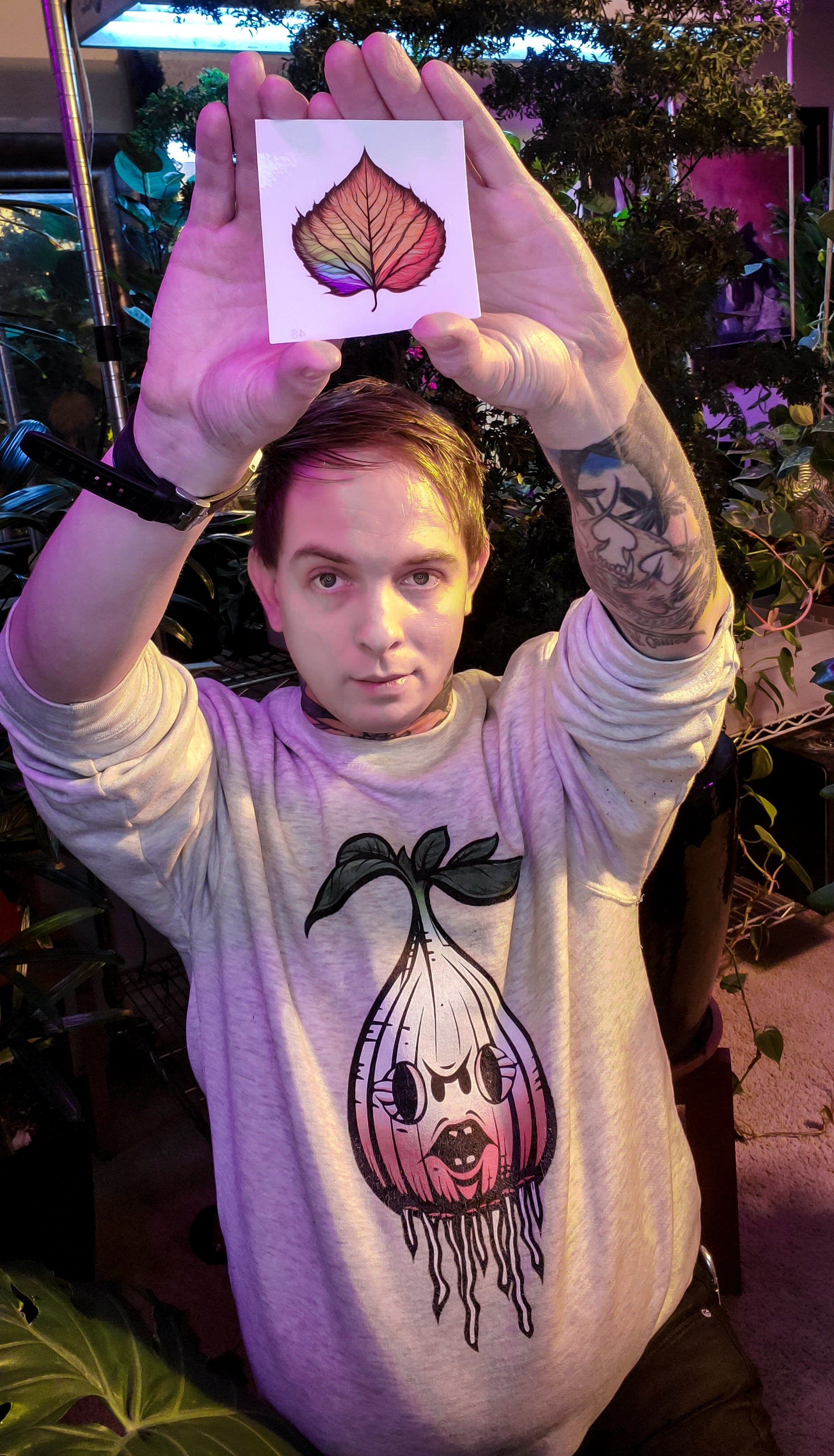 Individual with tattooed hands raises a colorful birch leaf sticker above their head.