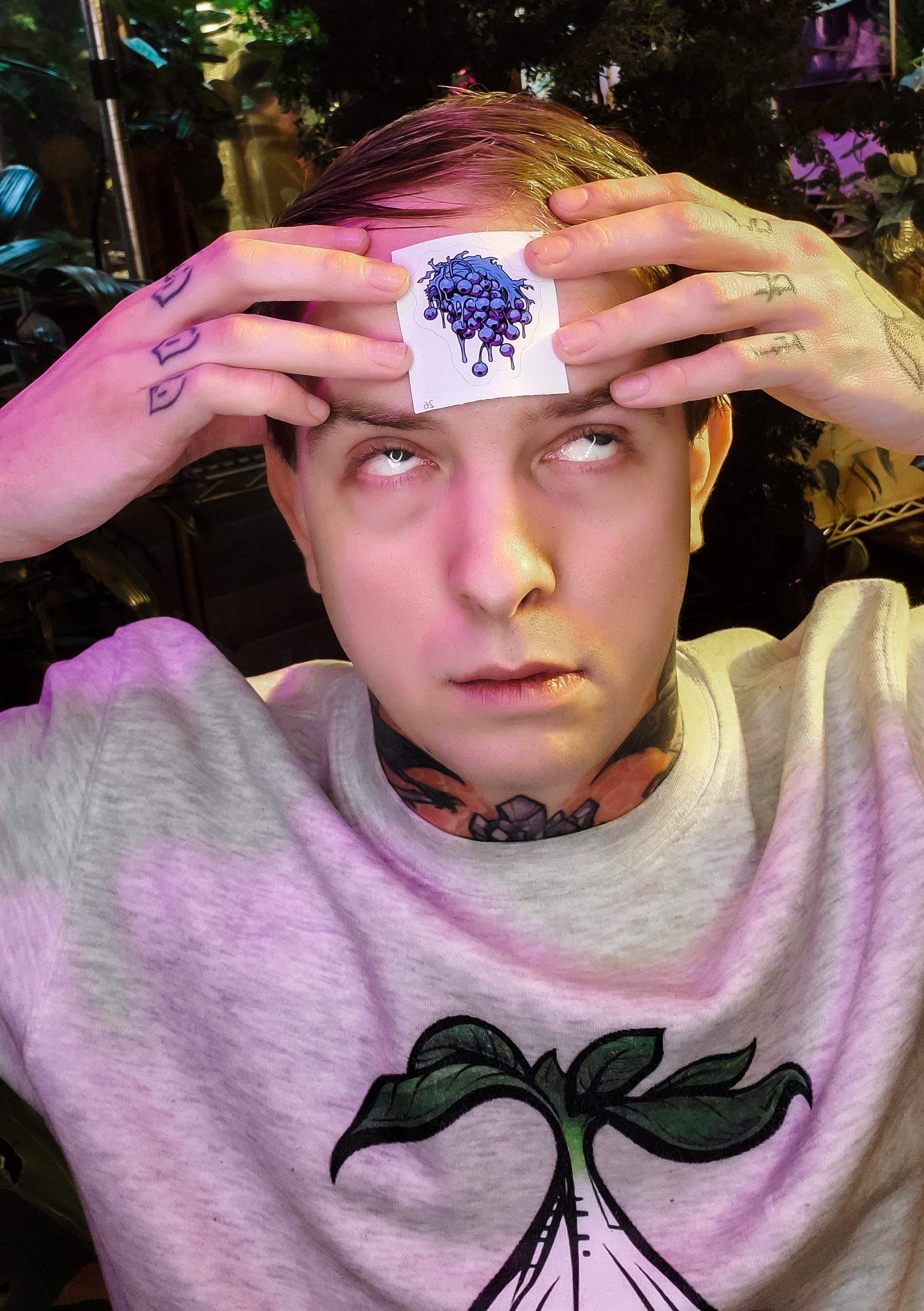 Individual places a horror-inspired blueberry cluster sticker on their forehead, tattoos visible