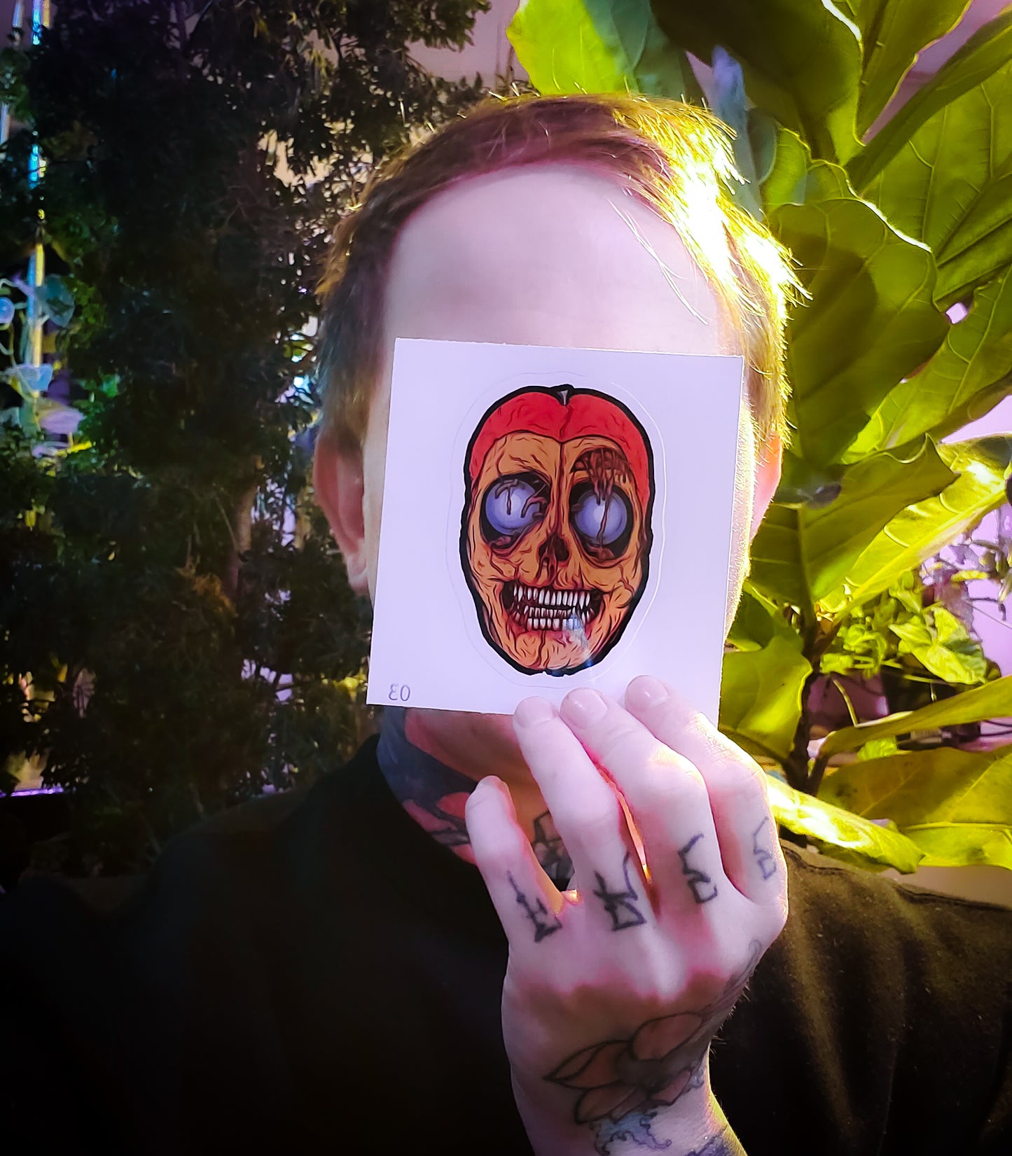 Person covering their face with a sticker of a spooky passion fruit skull, amidst foliage