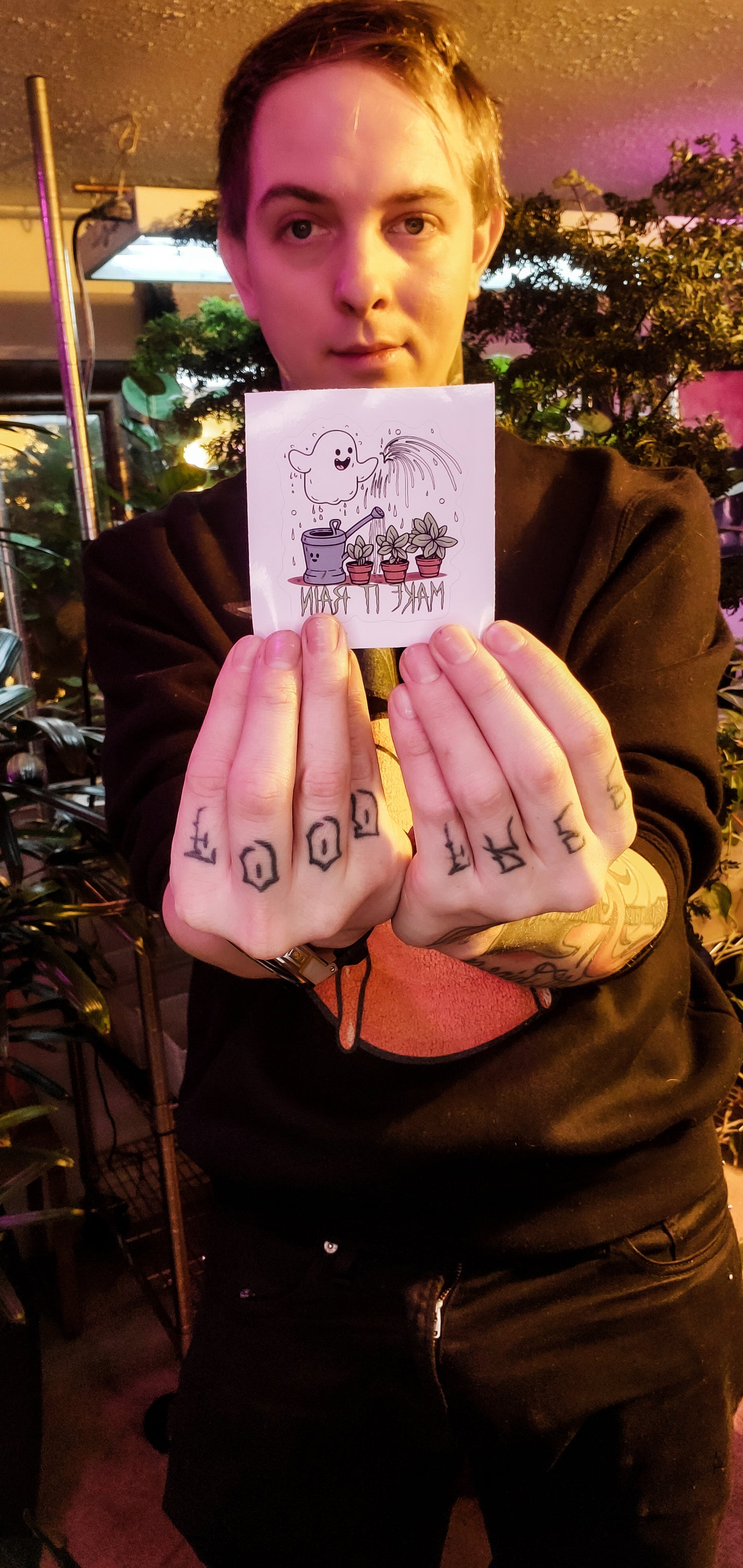 Person holds a playful ghost sticker with 'MAKE IT RAIN' text, houseplants in the background.