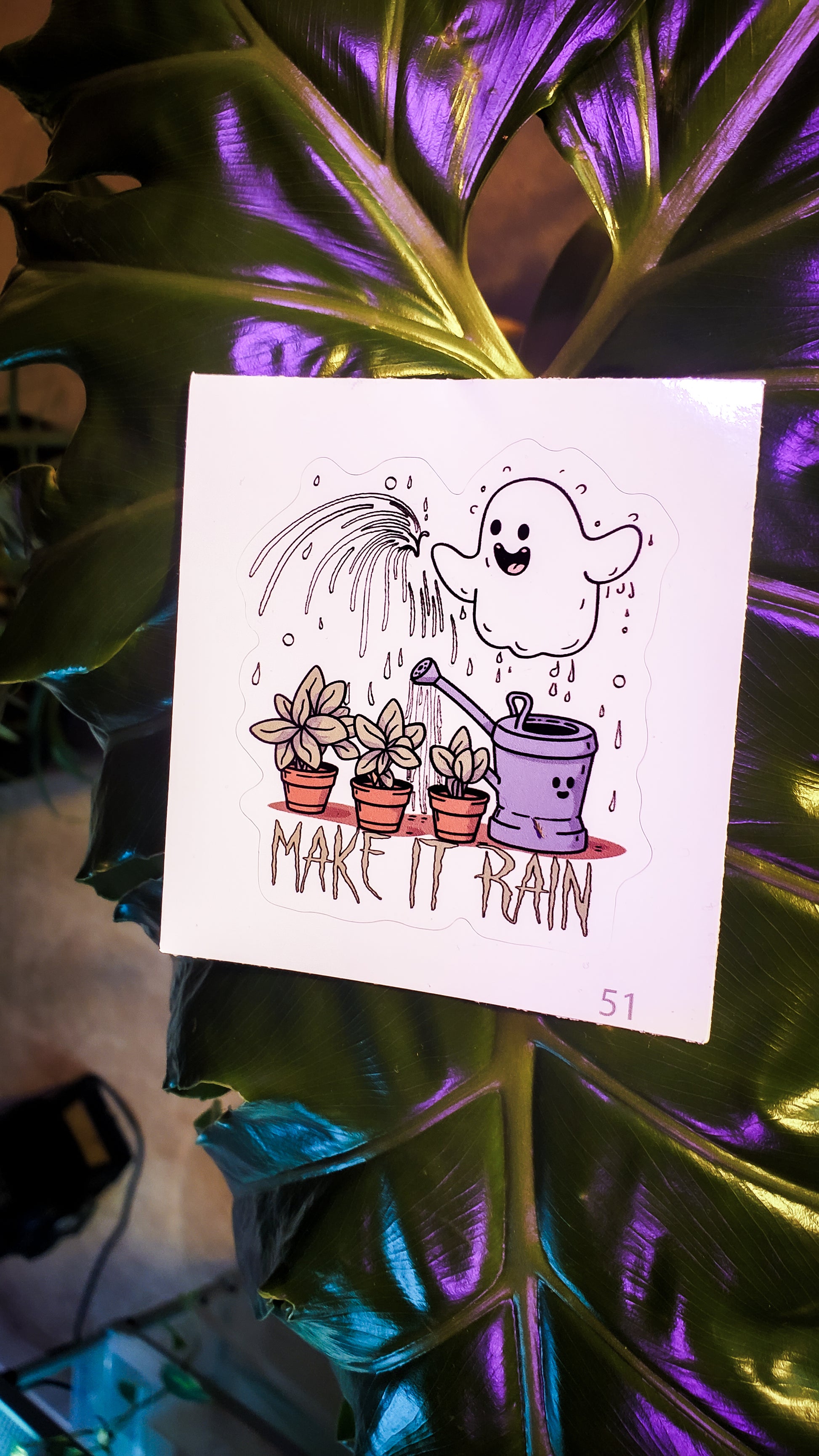 Cute ghost watering plants sticker with 'MAKE IT RAIN' phrase, set on a shiny leaf surface.