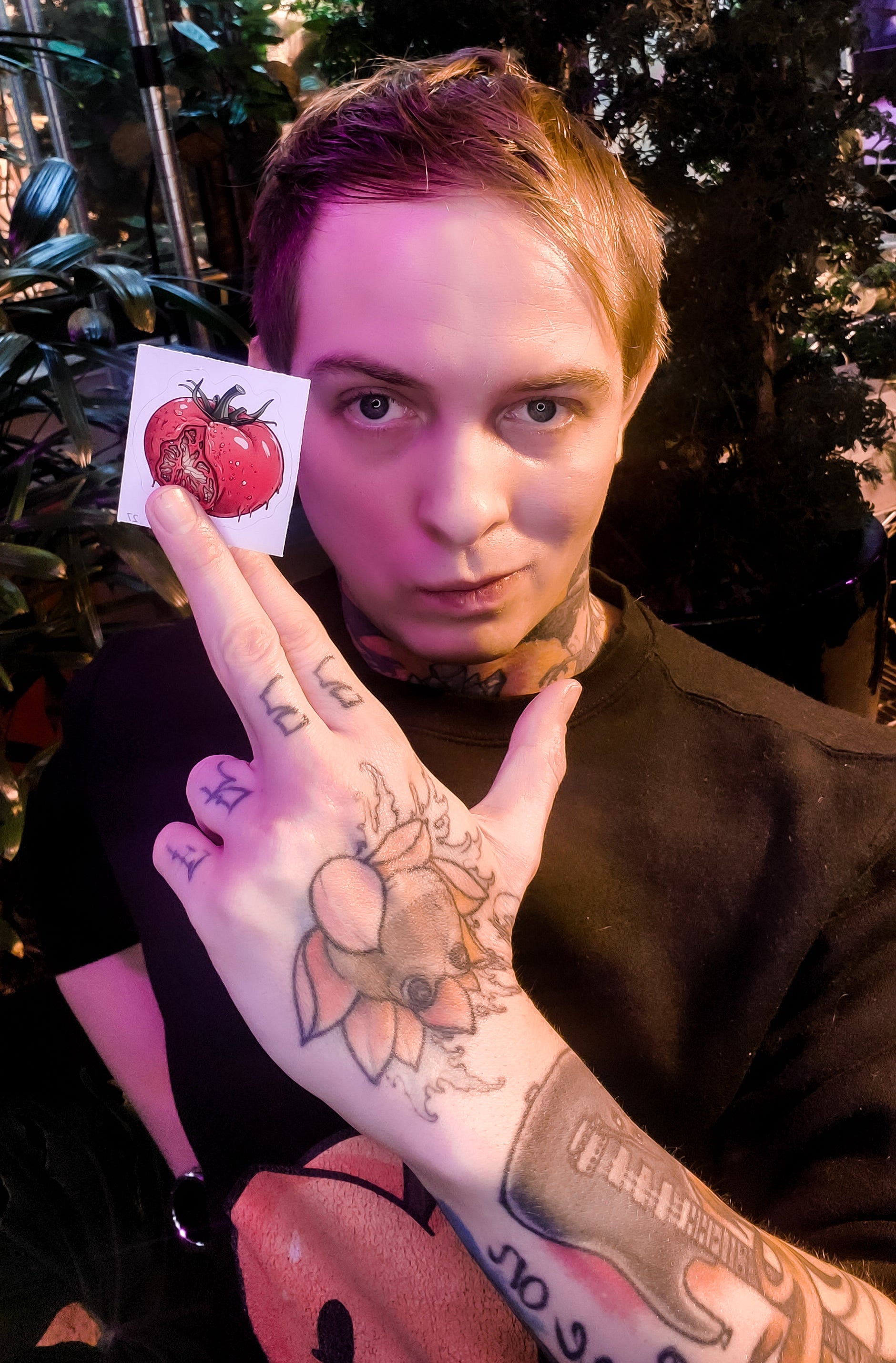 Person holding a tomato sticker with a horror twist over their eye, tattooed hand in focus.