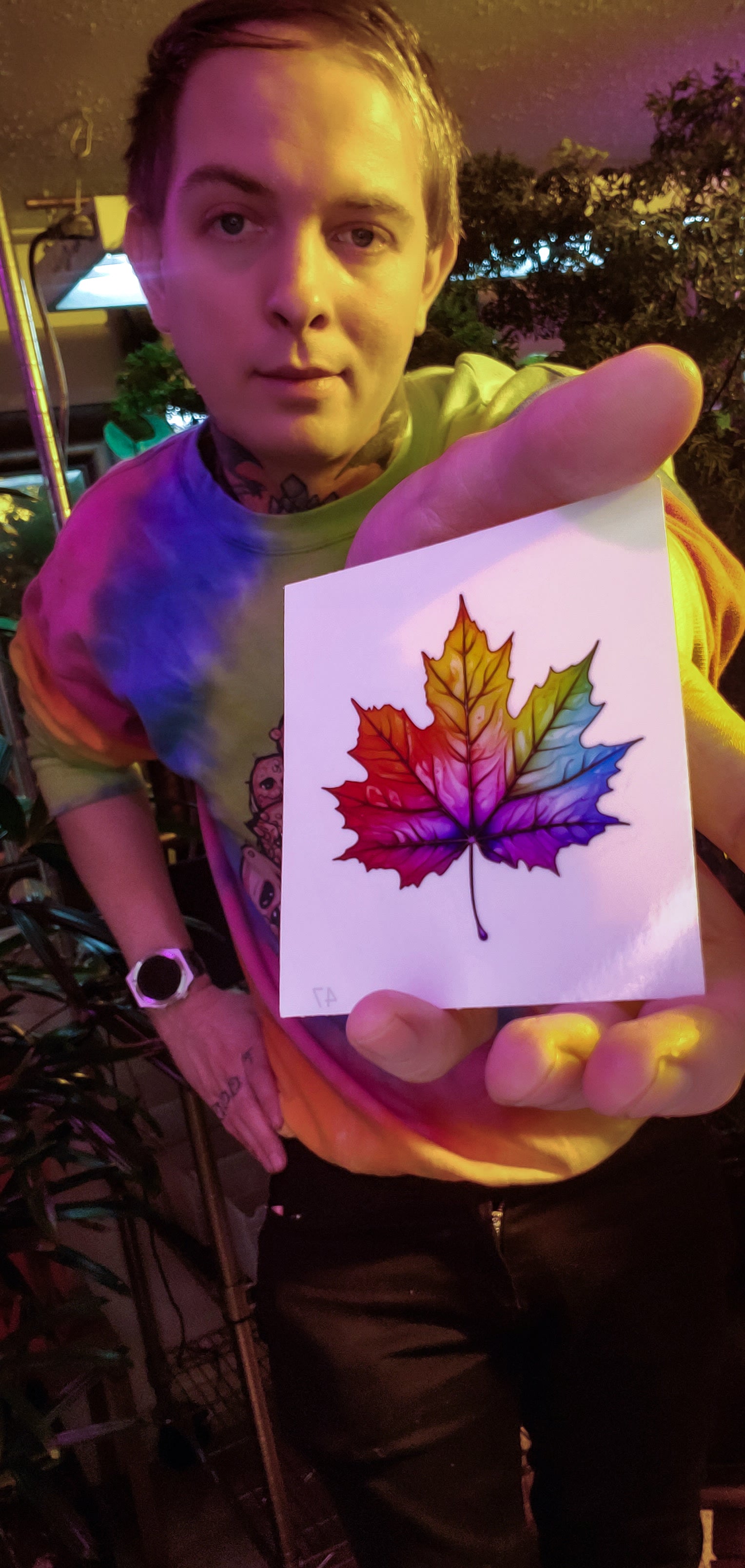 Person presenting a vibrant rainbow maple leaf sticker, with a tie-dye shirt and lush greenery behind