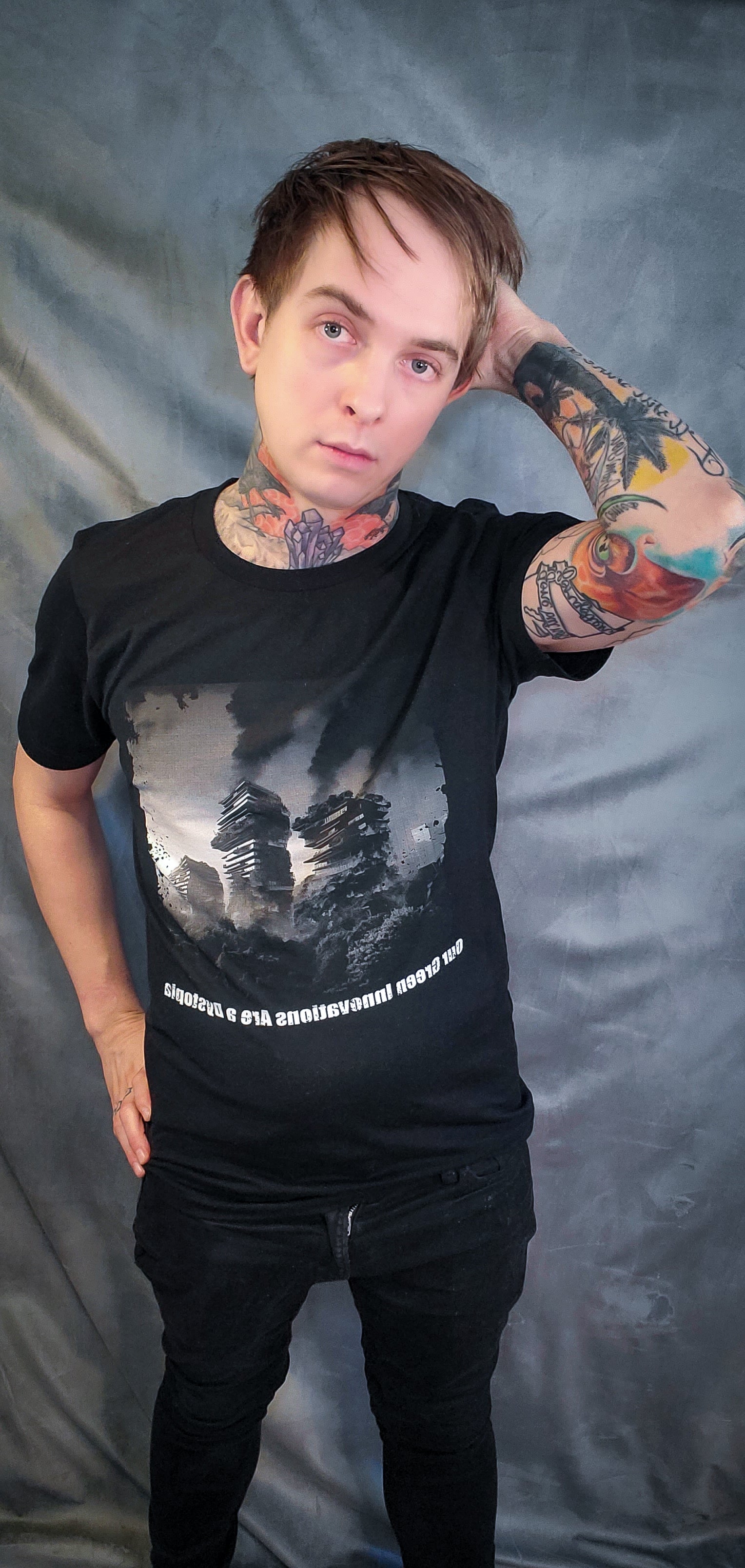 "Model showcases a punk rock black tee featuring a black and white album cover design with burning luxury high rises amid trees, and text 'Our green innovations are a dystopia.'"