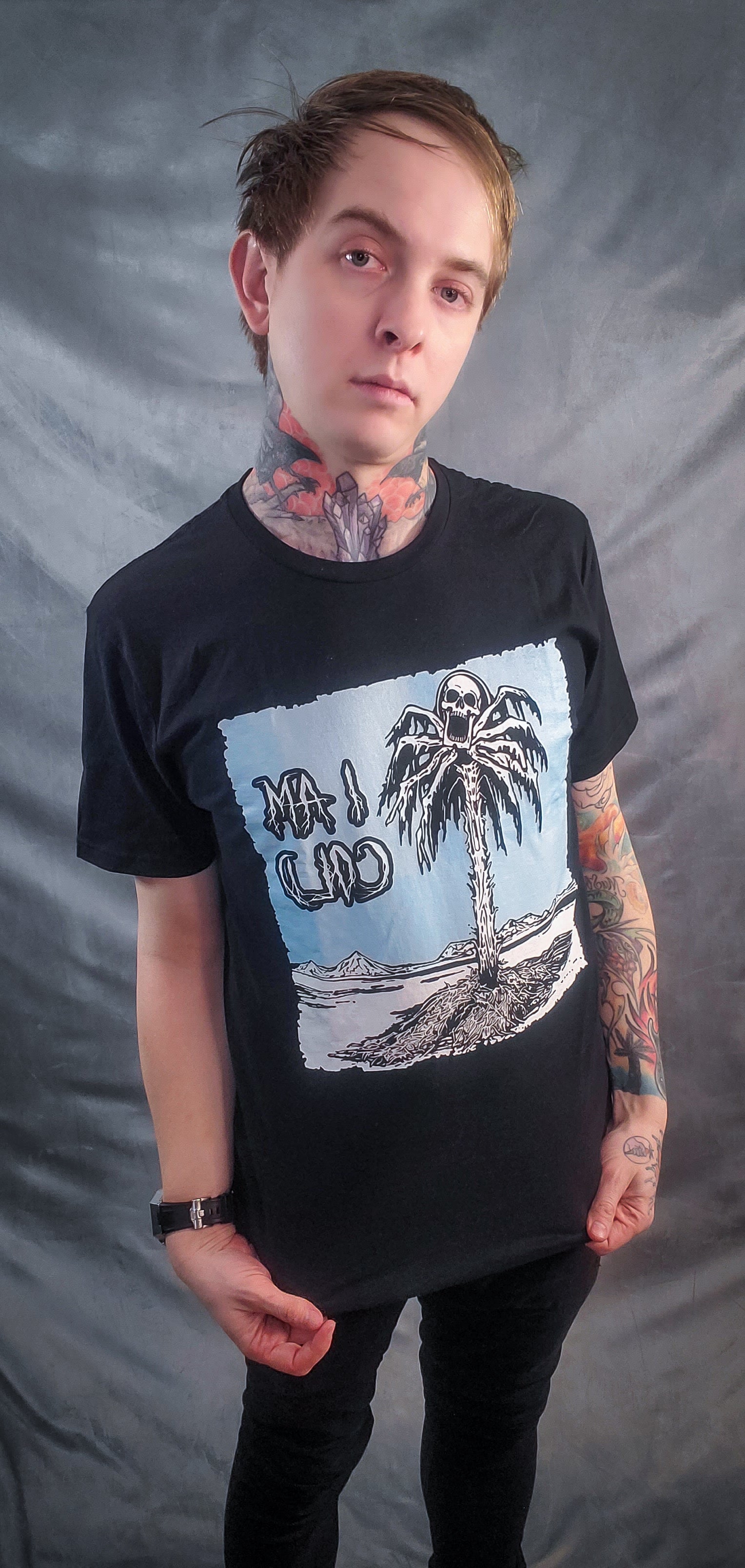 Tattooed person modeling a black t-shirt with a blue and white illustration of a palm tree in a frozen desert, accompanied by the bold text 'I am cold'.