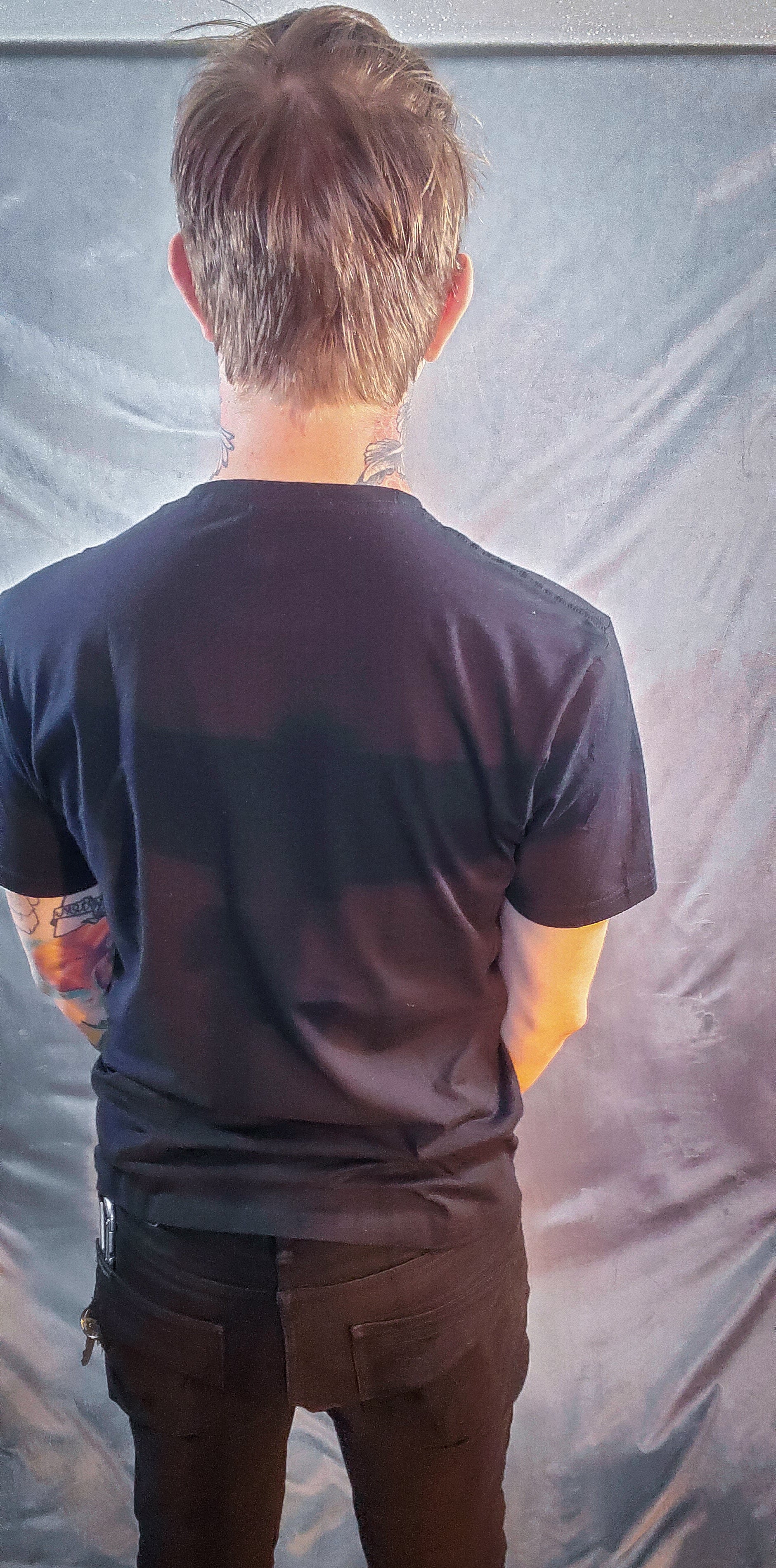 Back view of an individual in a black shirt and pants, facing a grey fabric background, hands on hips.