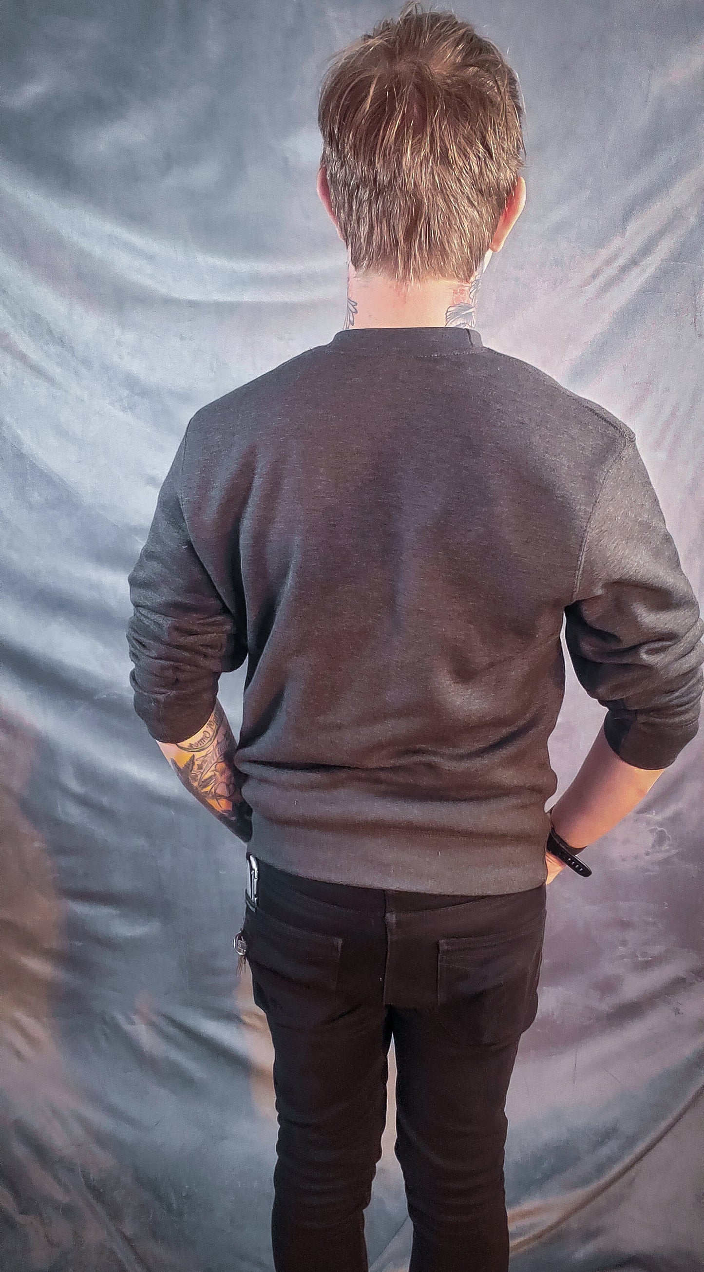 Back view of a dark sweatshirt worn by a person with tattooed arms, against a grey background.