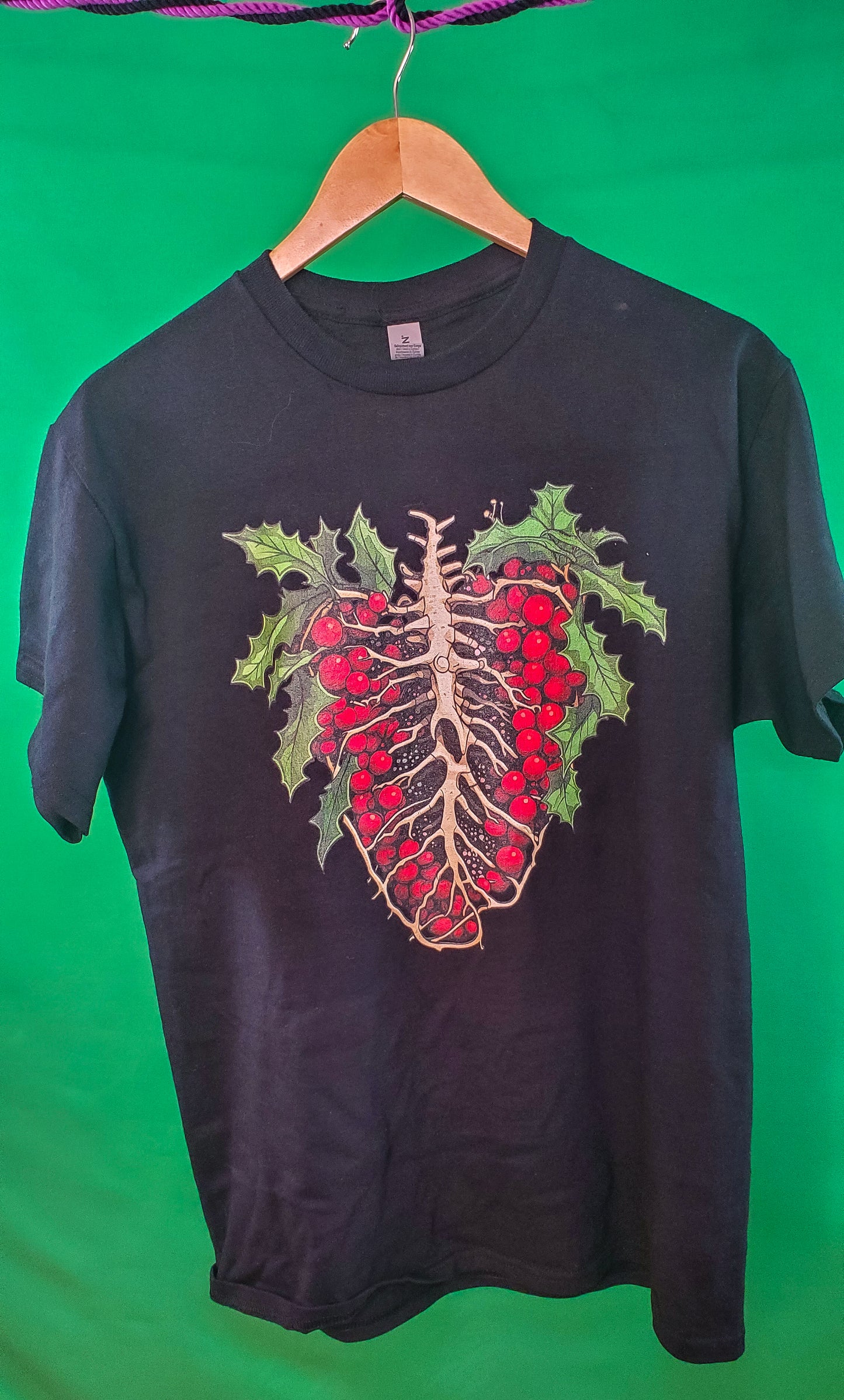 Black T-shirt with a ribcage and holly berries design, blending nature and anatomy, on a hanger with a green backdrop.