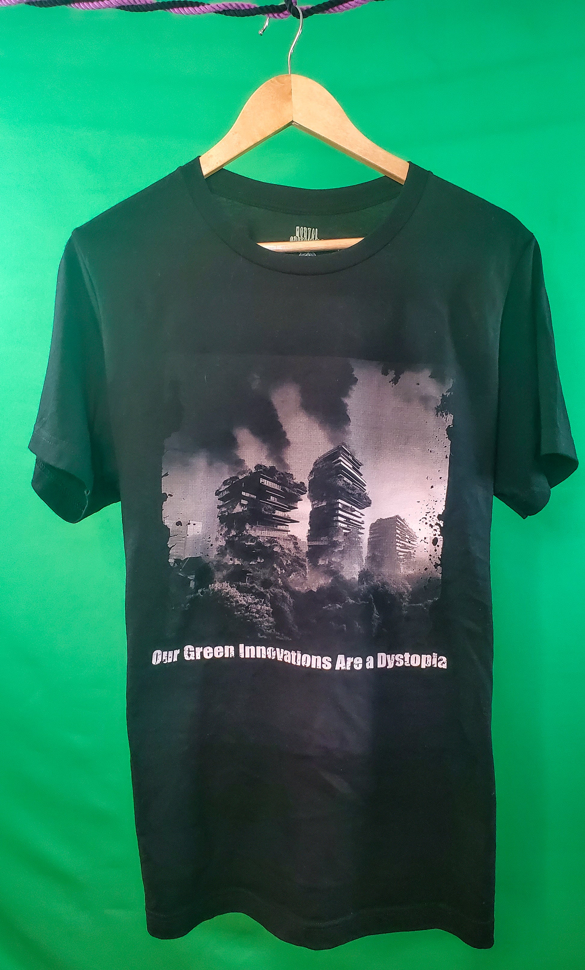 Black T-shirt with a dystopian design of burning skyscrapers overrun by foliage, titled 'Our Green Innovations Are a Dystopia. Against a green background.