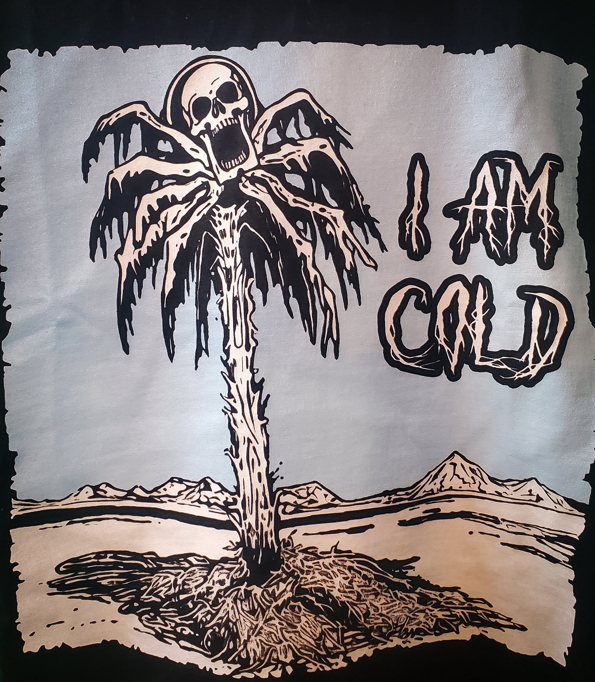 Close-up of a haunted frozen palm tree graphic with a skull on a T-shirt, set against a bleak desert background, with 'I AM COLD' text.