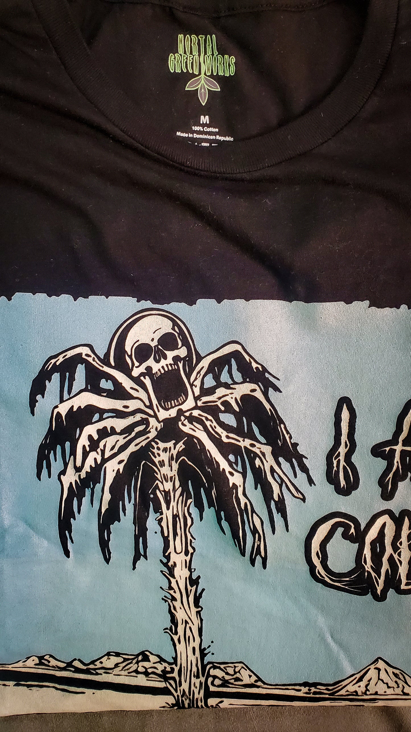 Mortal Greenworks logo on a T-shirt featuring a macabre frozen palm tree and skull design, embodying a cold desert horror theme.