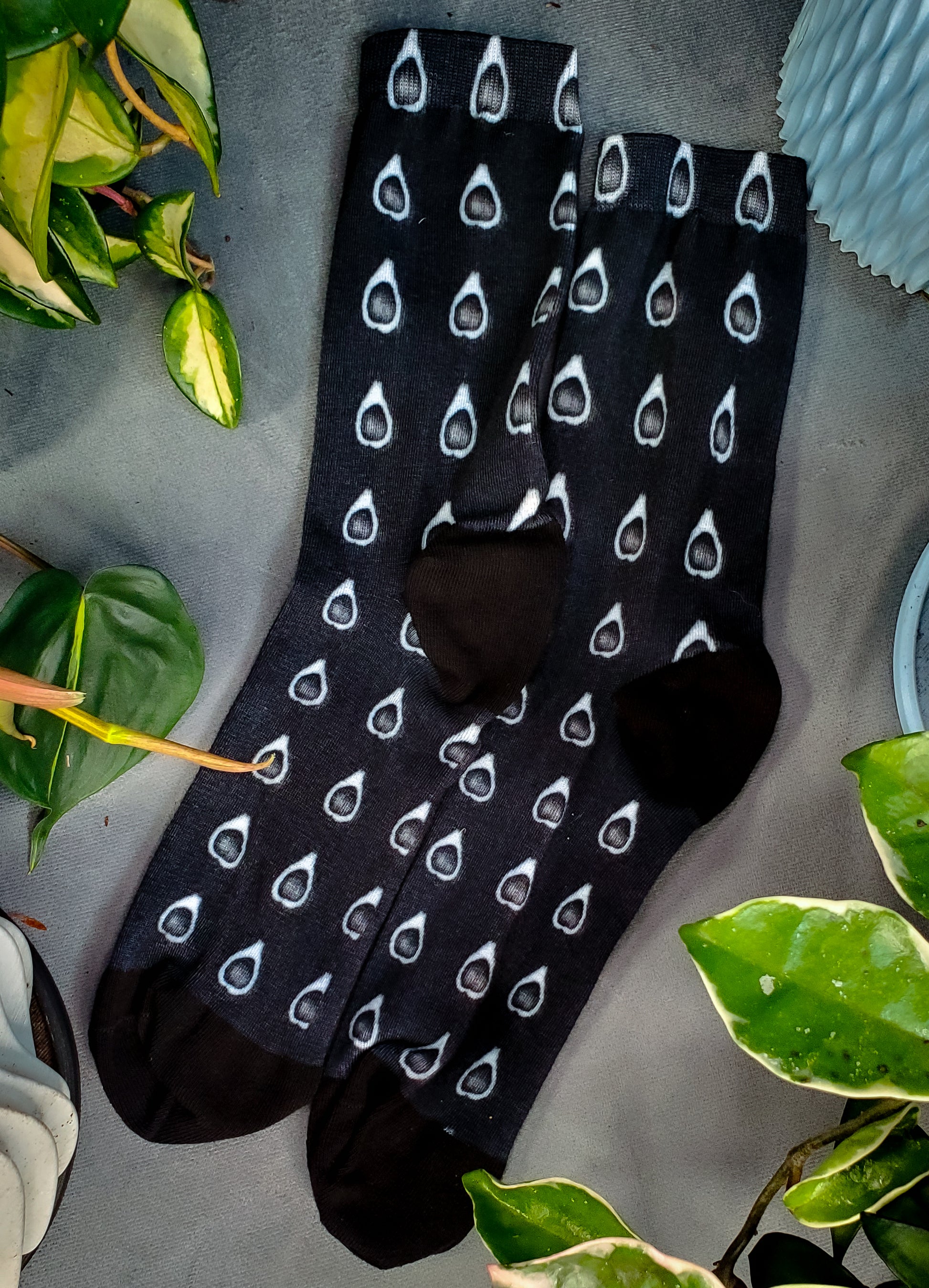 Comfortable socks with a unique black avocado horror print, nestled among vibrant houseplants, offering a quirky twist to plant-themed apparel.