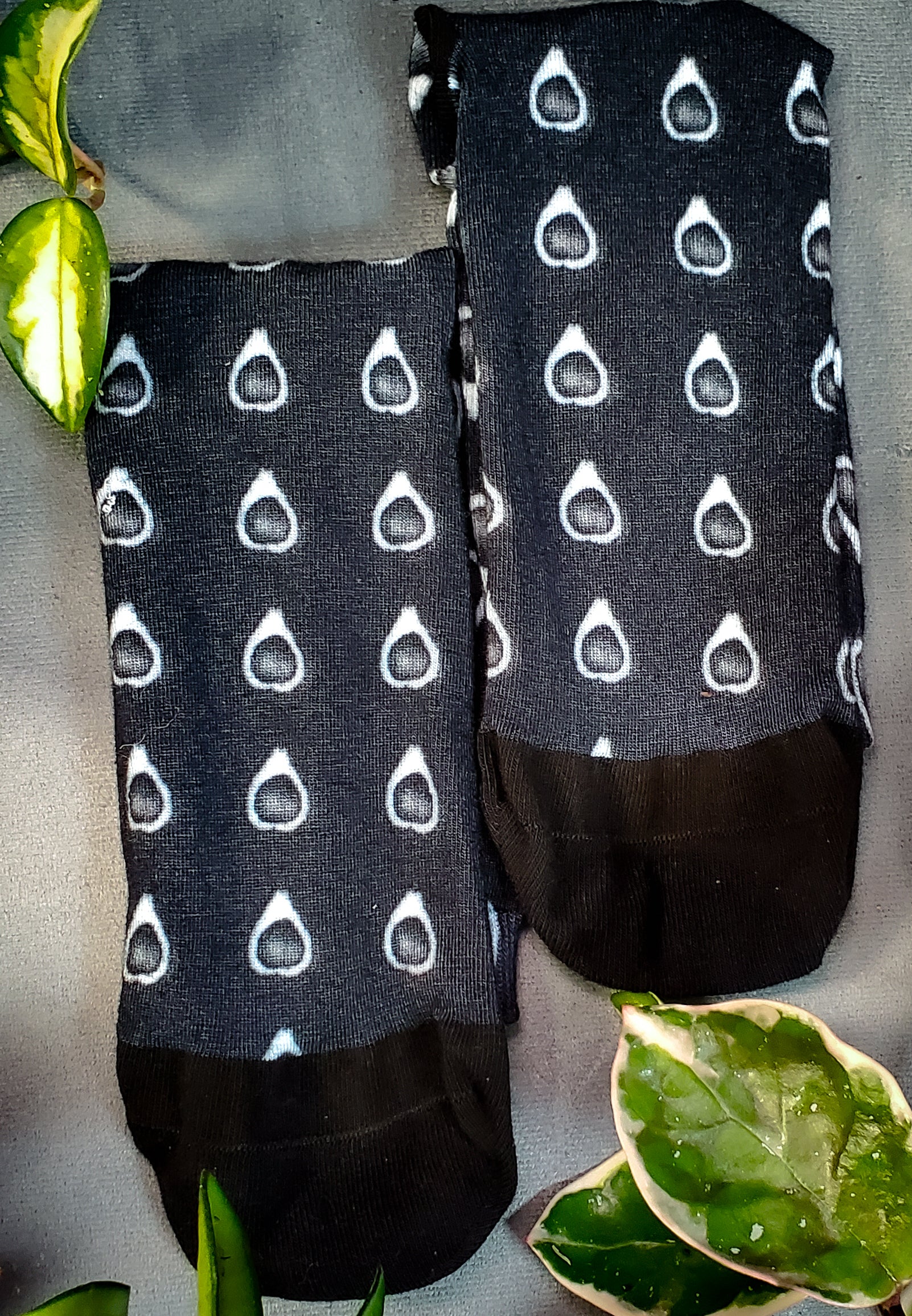 Flat-laid black socks featuring an avocado horror pattern, placed on a grey fabric surface, surrounded by greenery for a touch of natural charm.