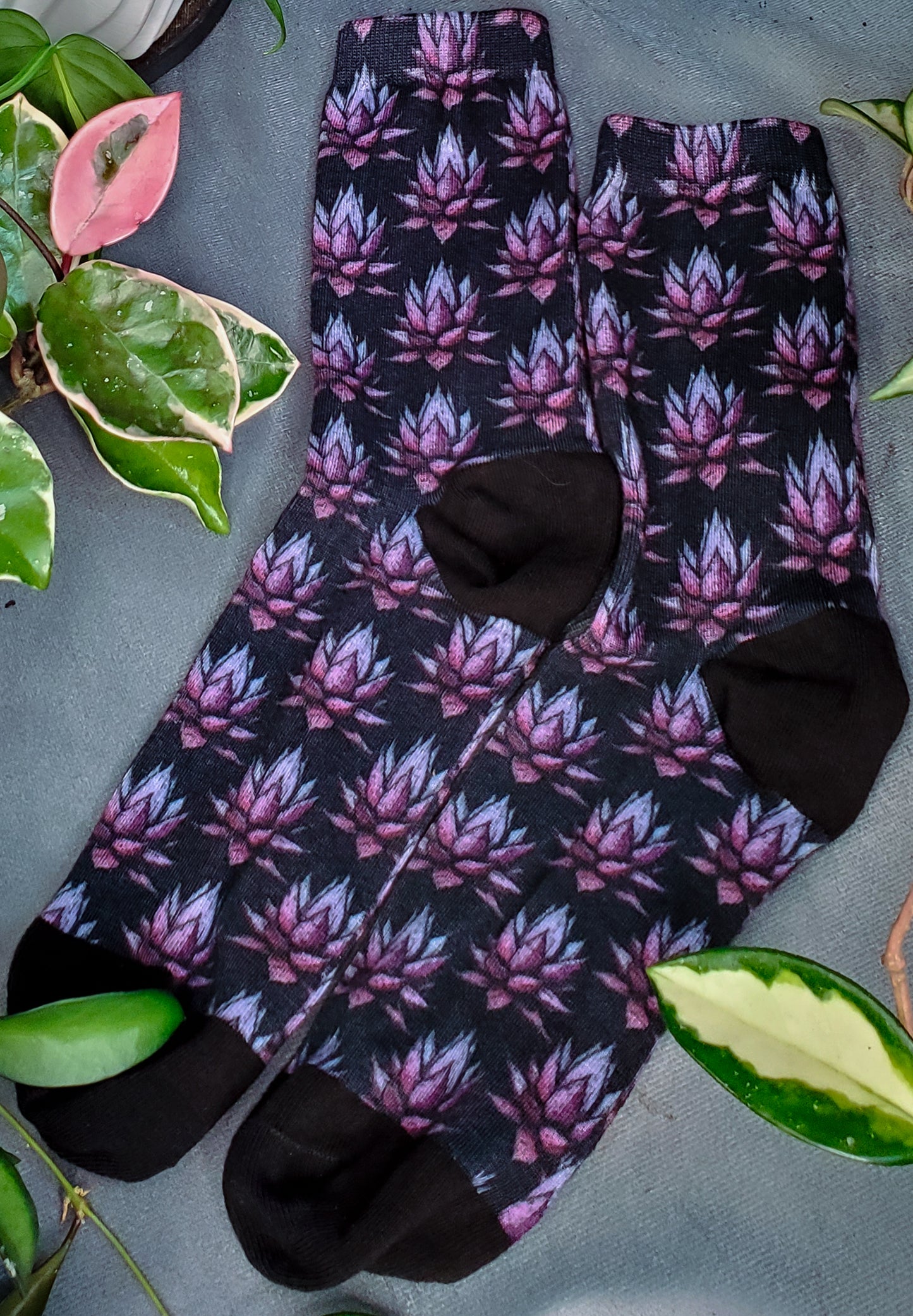 Socks with a dark, haunting purple agave pattern, surrounded by green and pink foliage on a grey surface.